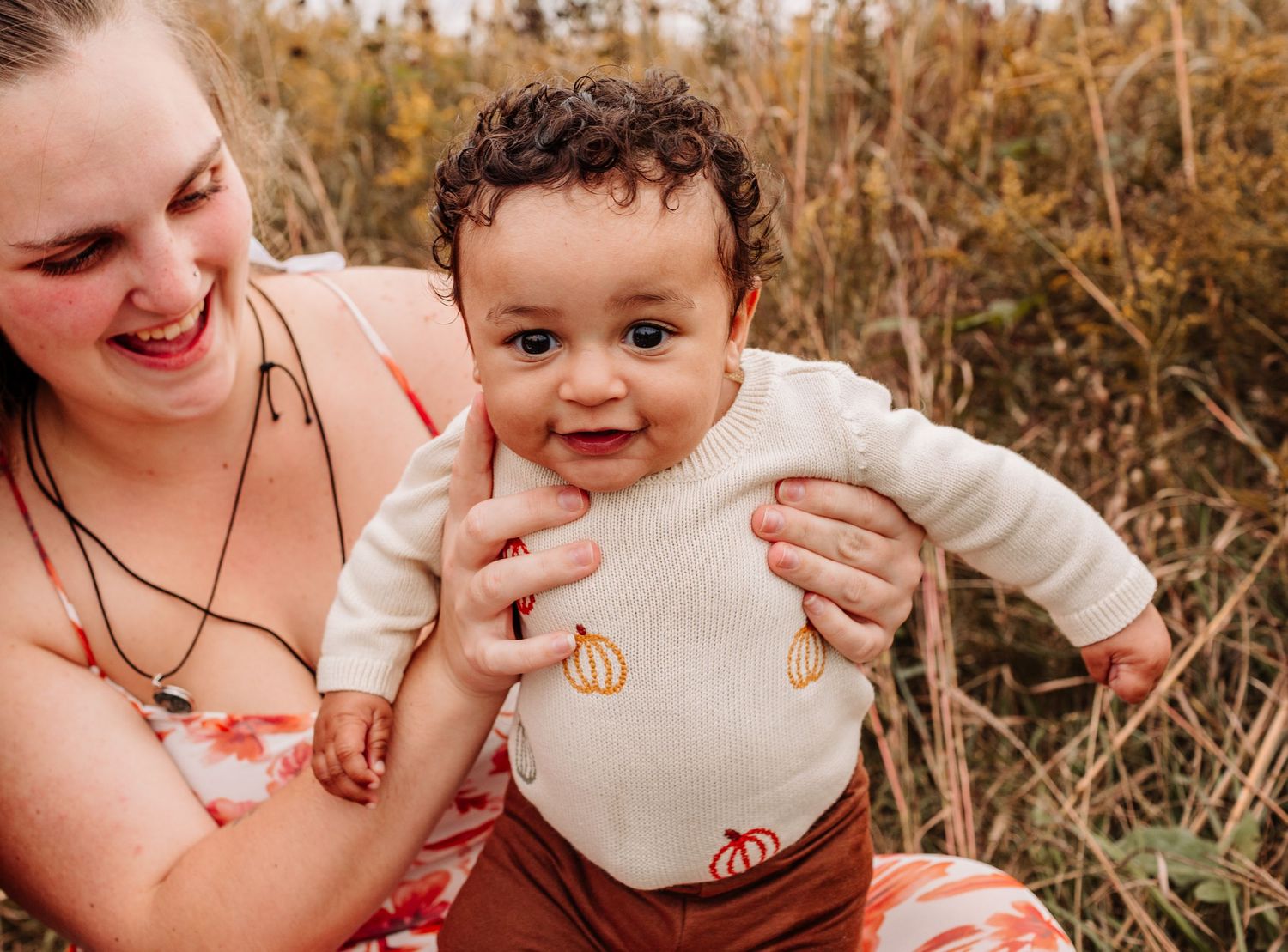A laughing mother holds her smiling baby in a cream pumpkin onesie outdoors in an autumn field.