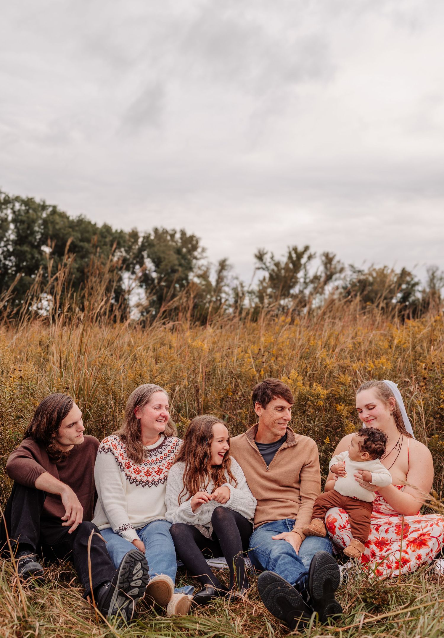 Family of five sitting together in an autumn field, smiling and holding a newborn baby.