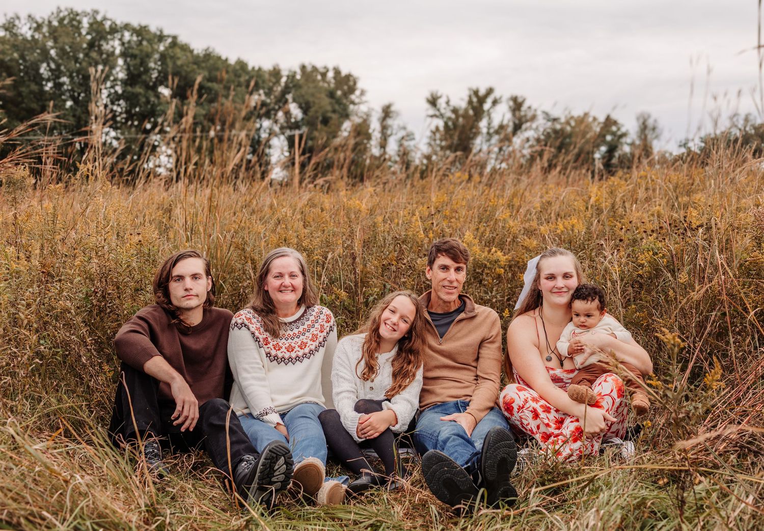 Family of six sitting together in a golden autumn field, smiling warmly for a fall outdoor portrait.