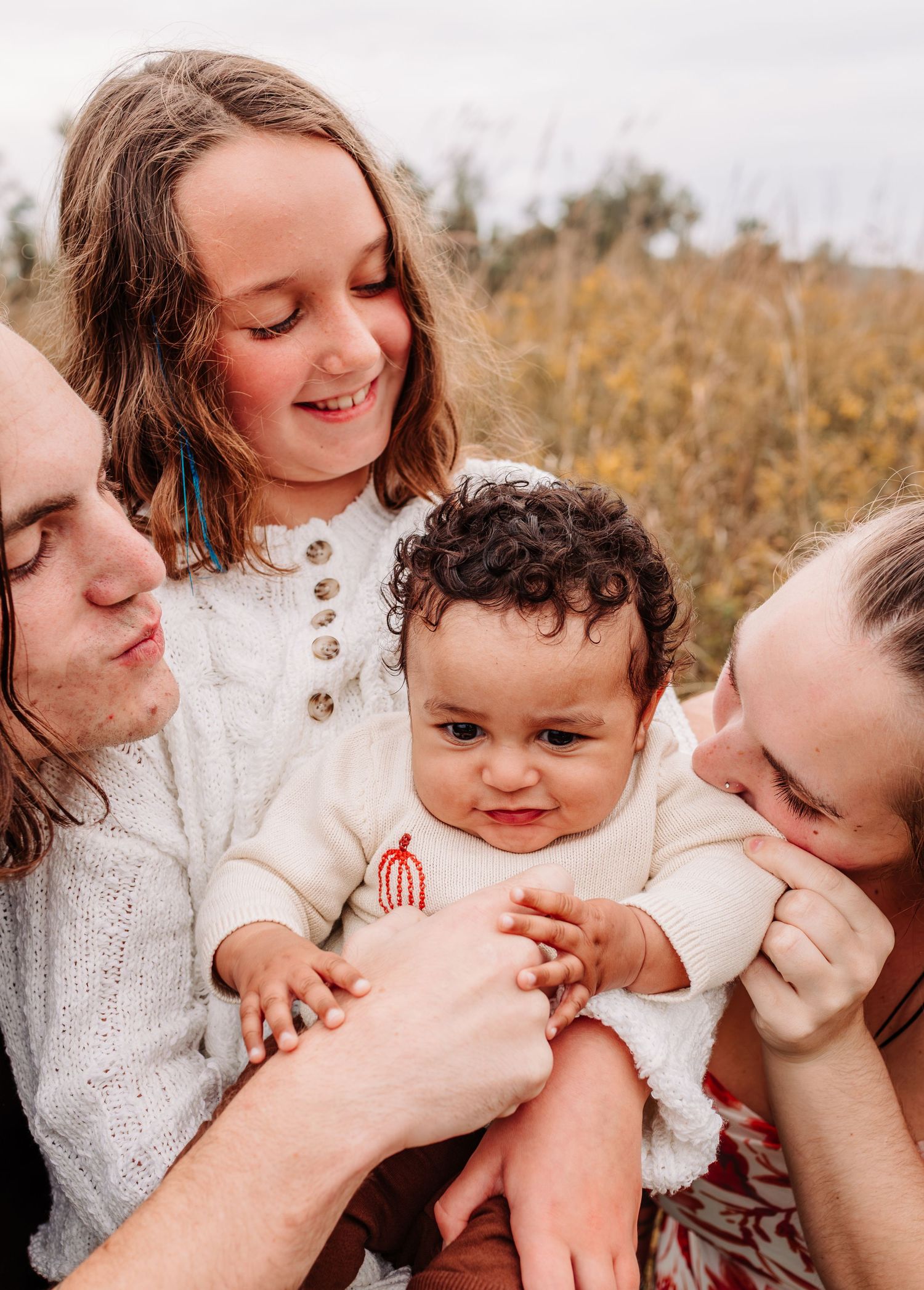 A joyful family with two adults, a young girl, and a curly-haired baby enjoying an outdoor moment together.