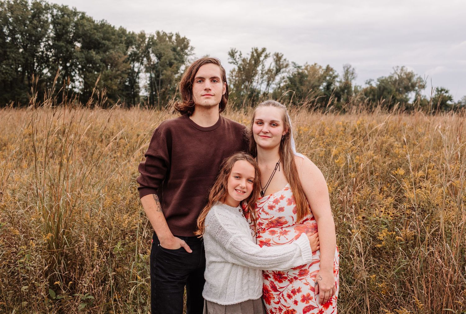 Three siblings pose together in a golden field, with tall grass and trees in the background on a cloudy day.