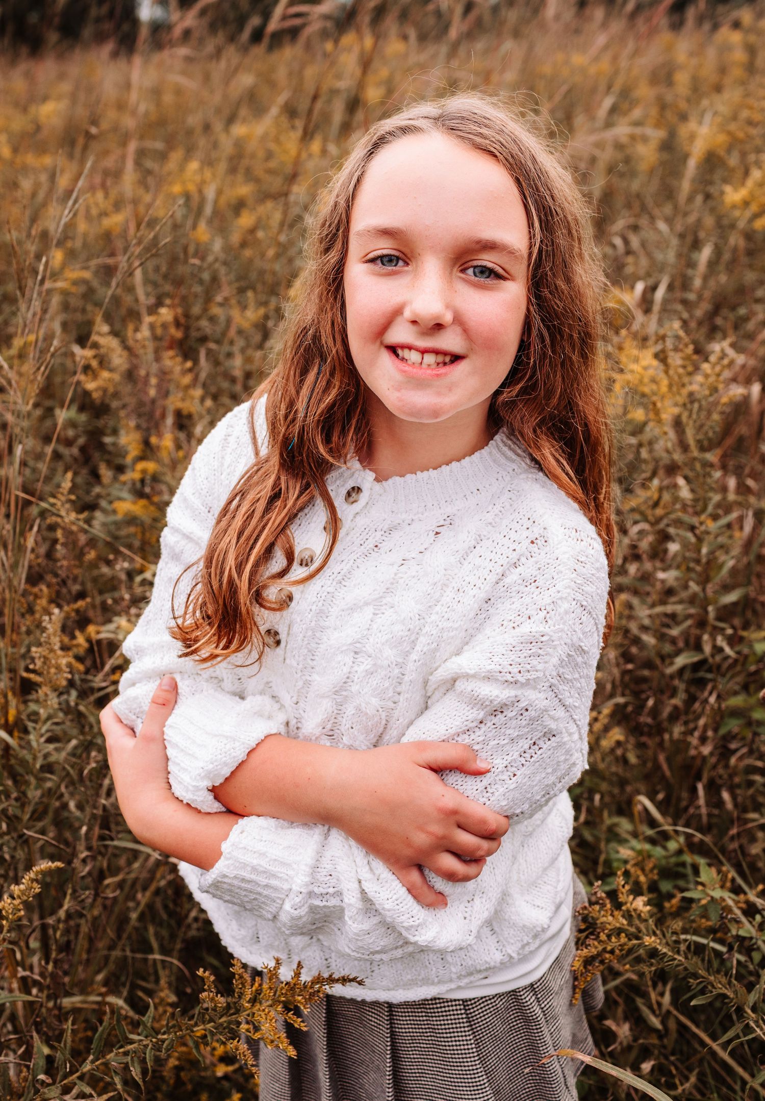 Smiling girl in white cable-knit sweater posing in autumn field with golden wildflowers.