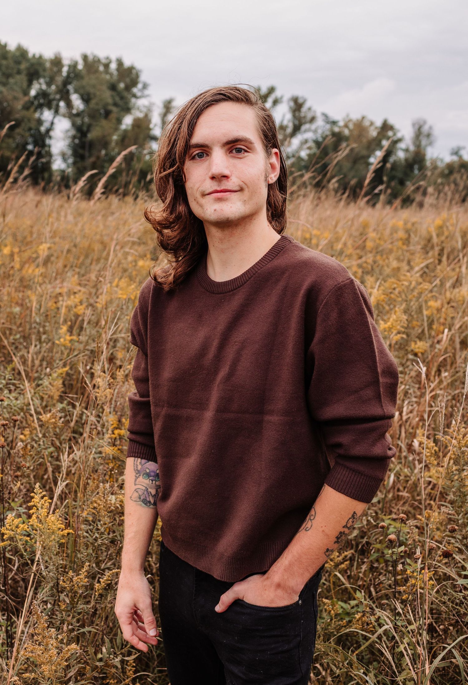 Young man with long hair and tattoos wearing a burgundy sweater standing in a golden field.