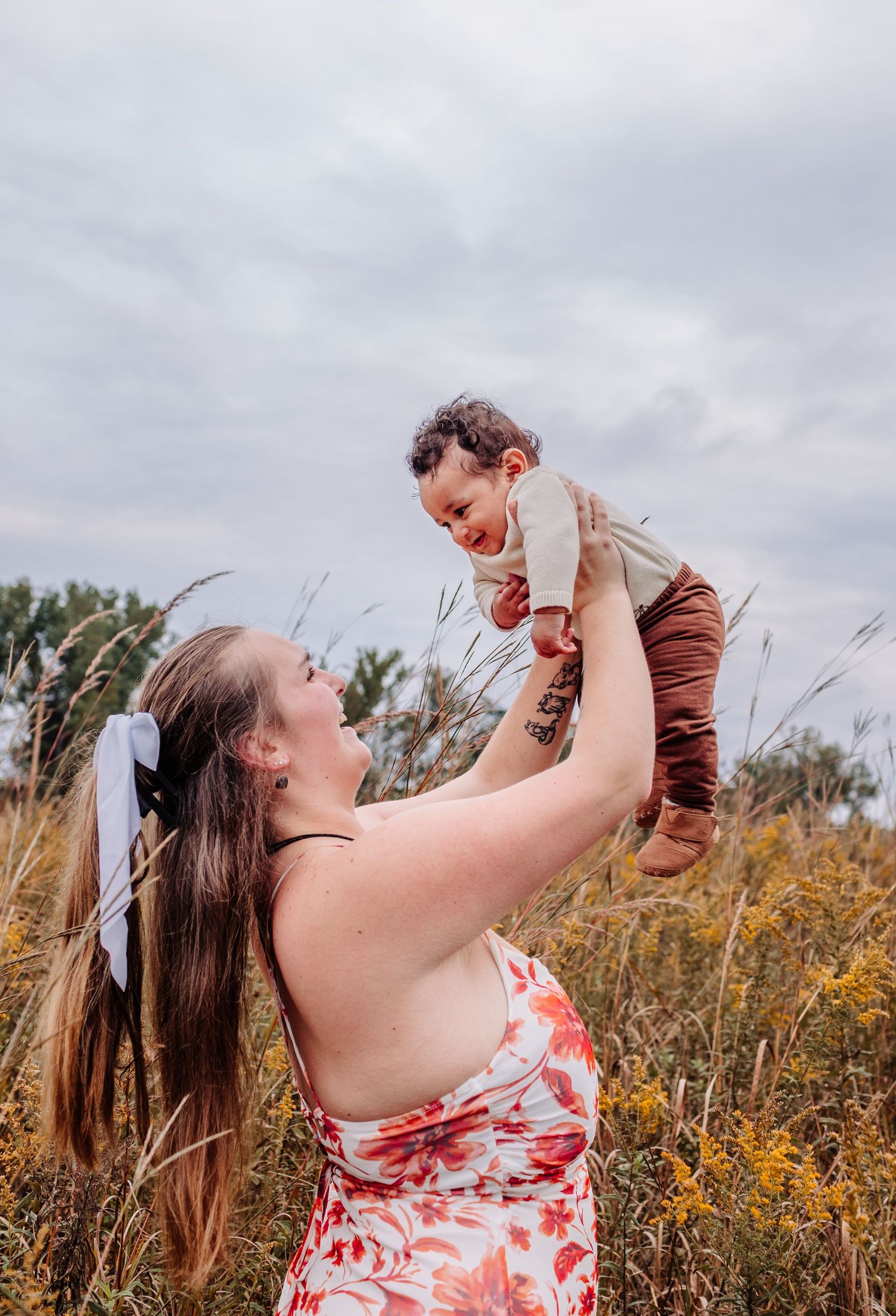 A mother lifts her laughing baby in a golden autumn field while wearing a floral dress with a white bow in her hair.