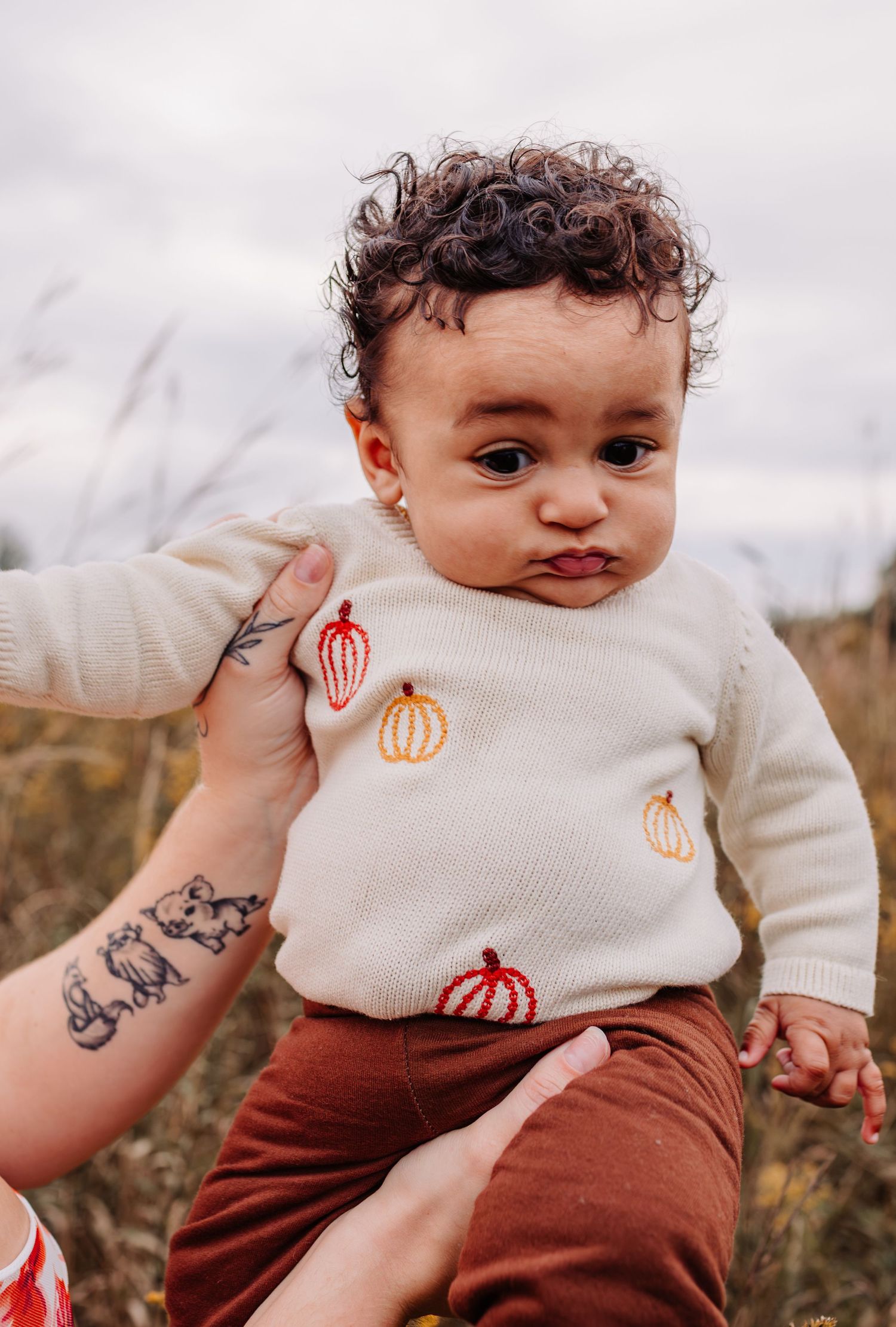 An adorable baby with curly hair wears a cream pumpkin-embroidered sweater and rust pants in an autumn outdoor setting.