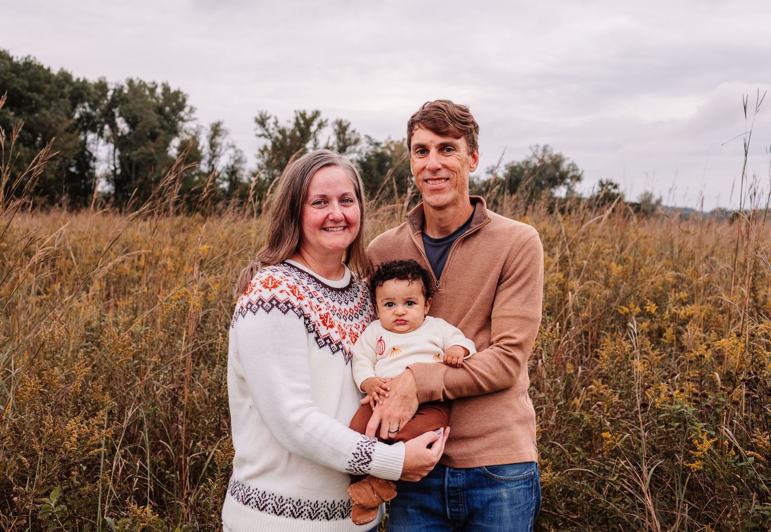 Family portrait of two adults holding a baby in a golden field with trees in the background.