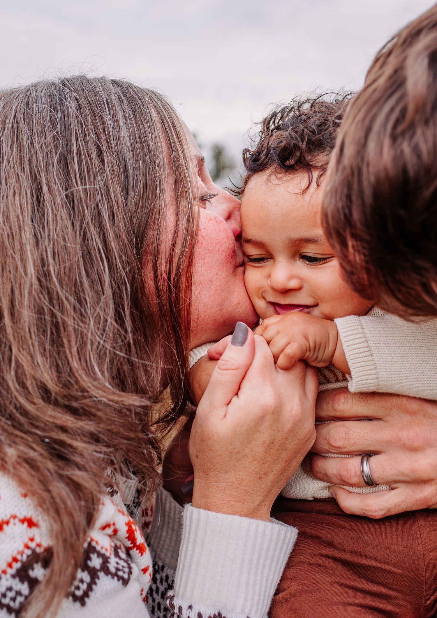 Two parents lovingly kissing their smiling curly-haired baby on the cheeks in a tender family moment.