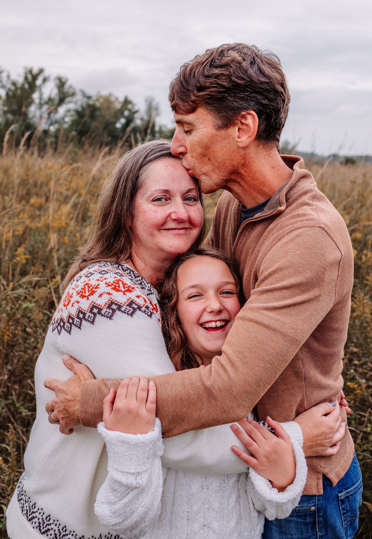 Family of three sharing warm embrace in golden autumn field, parents hugging smiling daughter.