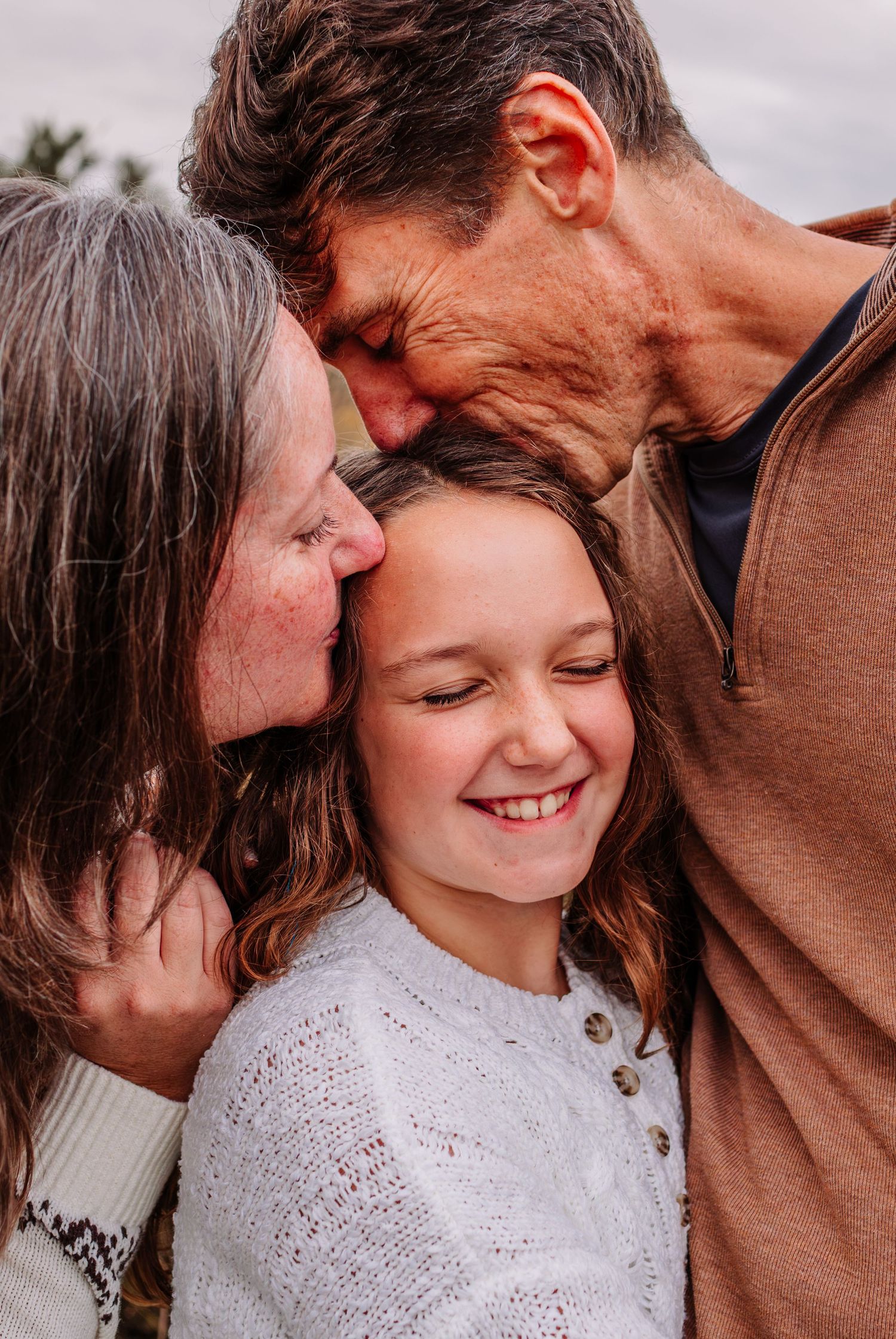 Two grandparents lovingly kissing a smiling young girl on the cheeks in a warm family portrait outdoors.