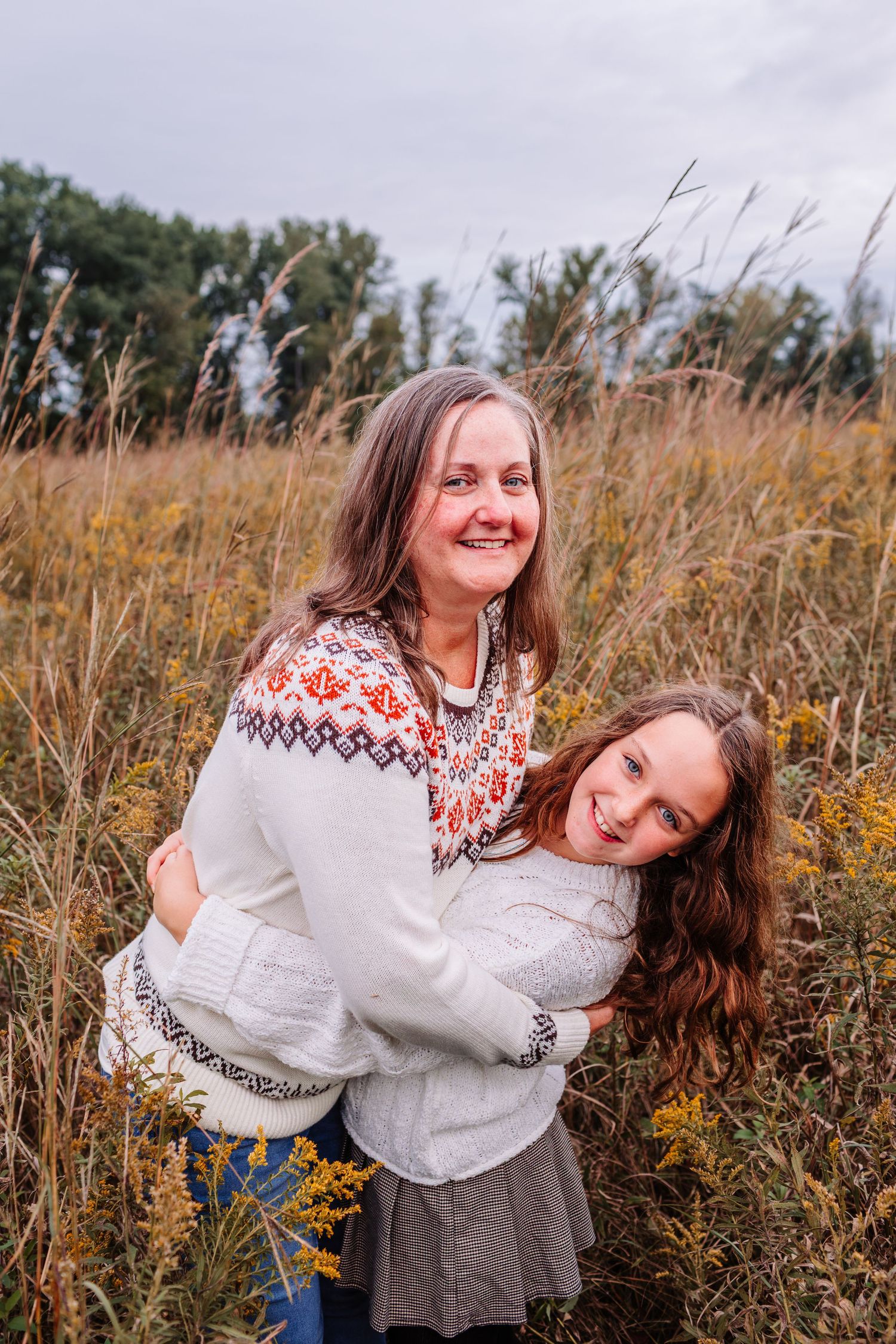 A woman and young girl in matching Nordic sweaters embrace warmly in an autumn field of golden wildflowers.