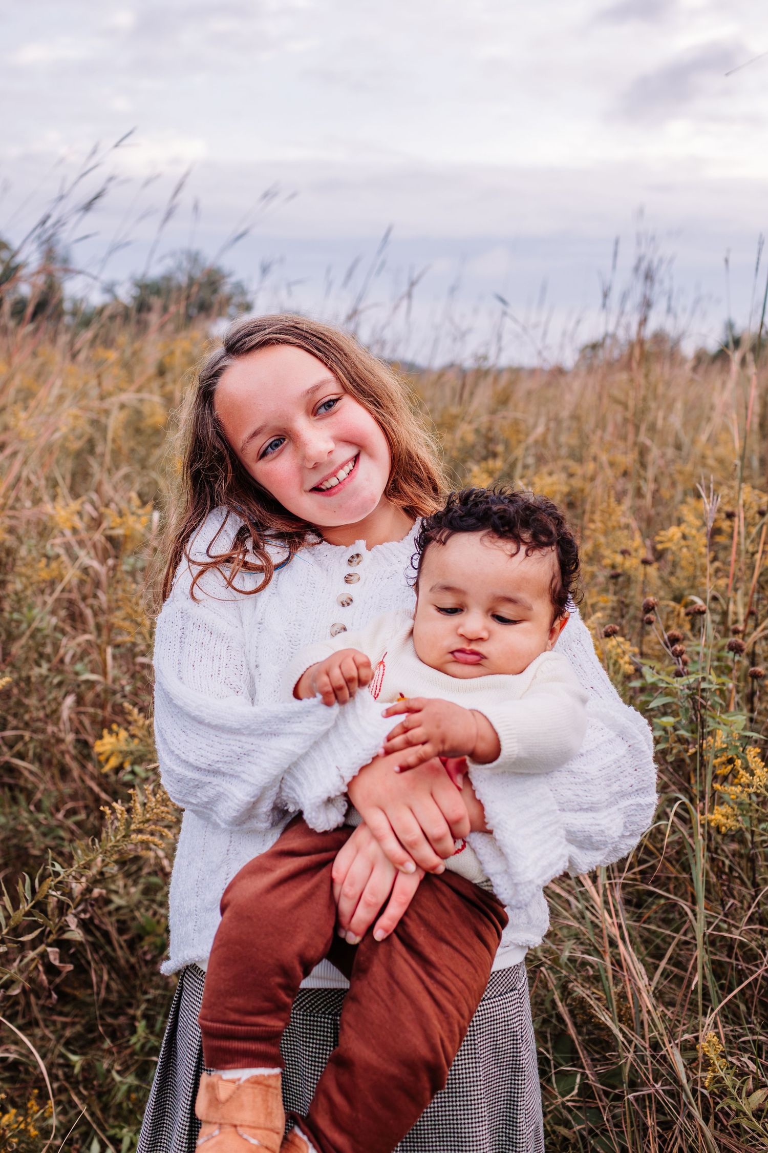 A smiling girl in a white sweater holds a sleepy baby in rust pants in a golden autumn meadow.