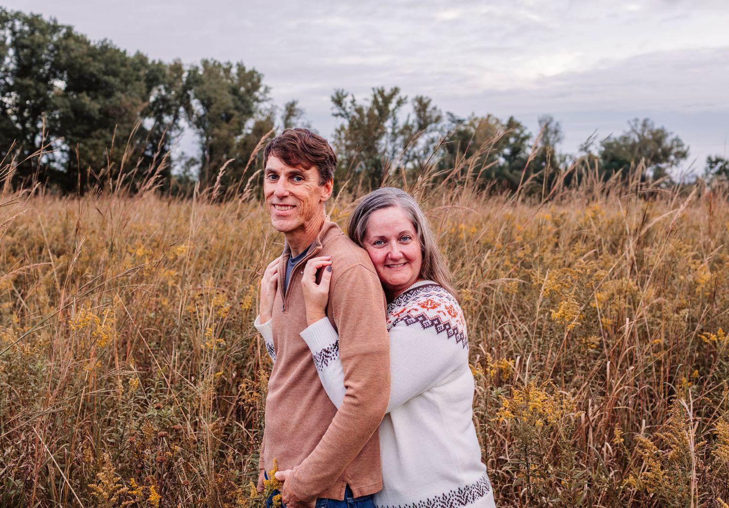 Two people smiling back-to-back in a golden field with trees under a cloudy sky.