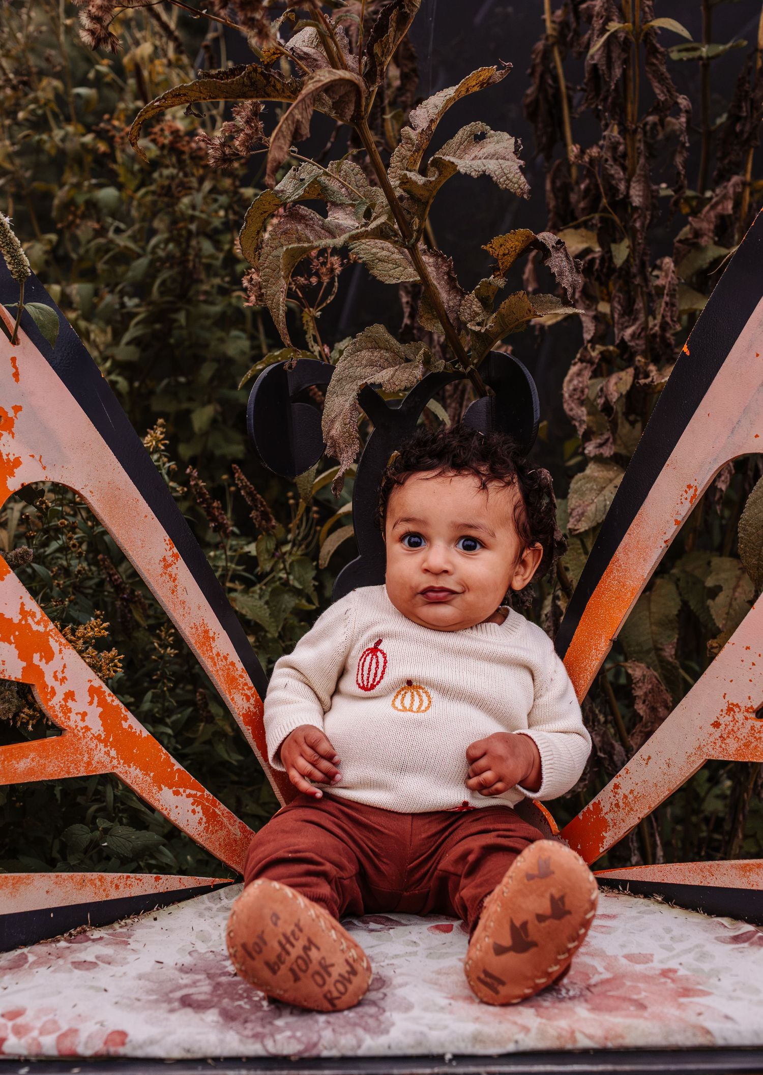 Adorable baby in a pumpkin sweater sitting in a rustic orange metal chair outdoors.