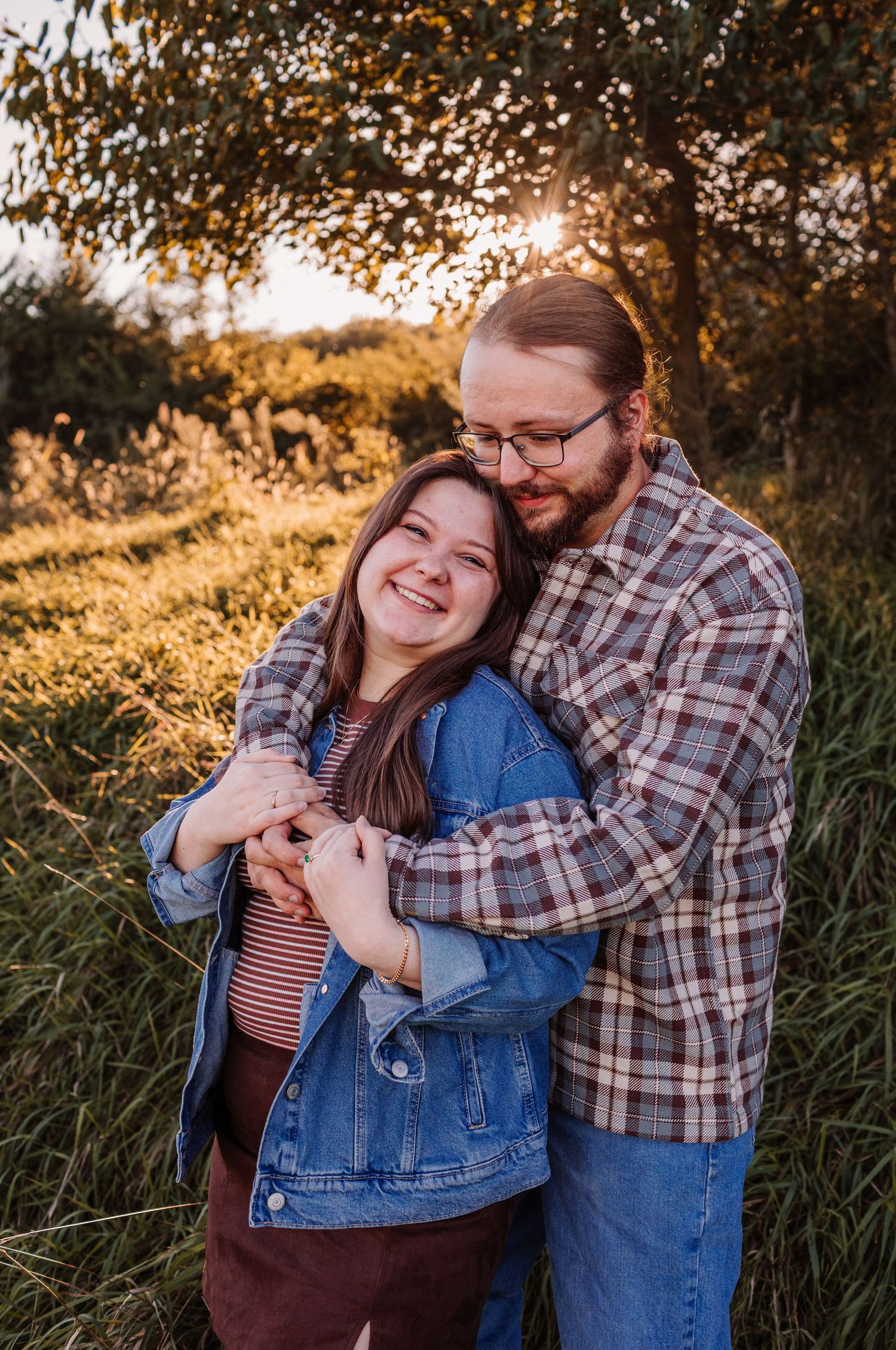 Couple embracing outdoors at golden hour, woman smiling in denim jacket, man in plaid shirt, surrounded by lush greenery.
