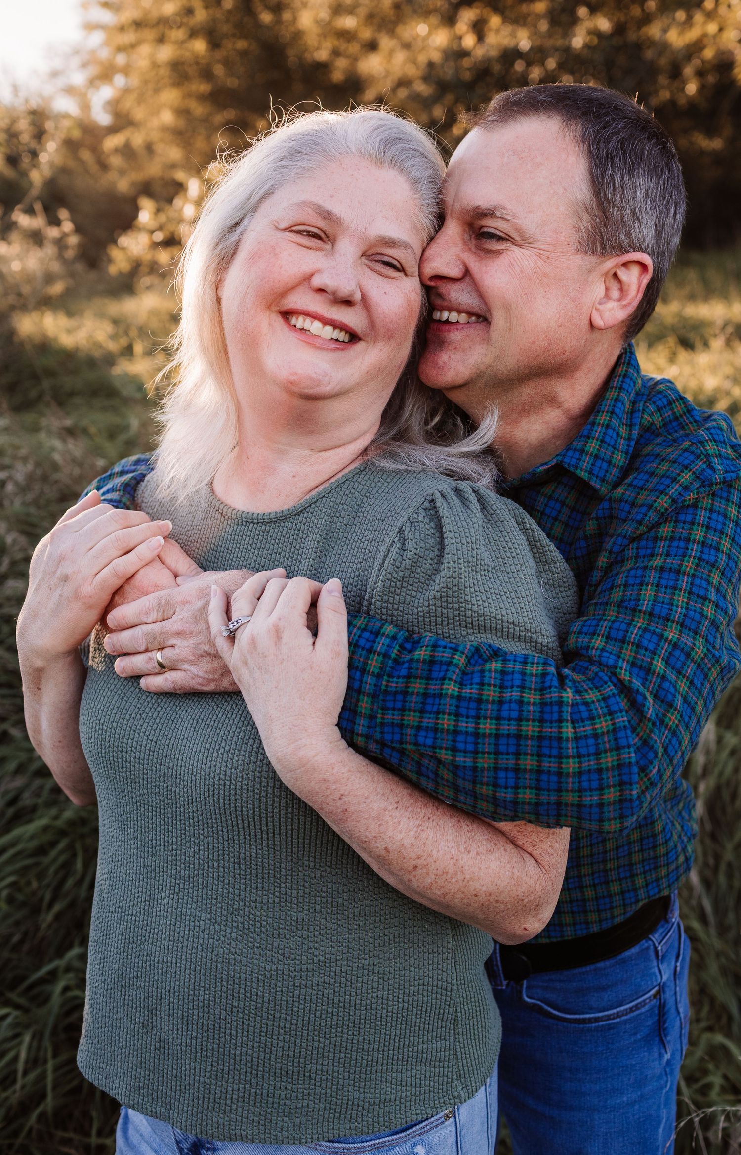 A happy older couple embracing outdoors, woman with silver hair in green top, man in plaid shirt, smiling warmly at golden hour.