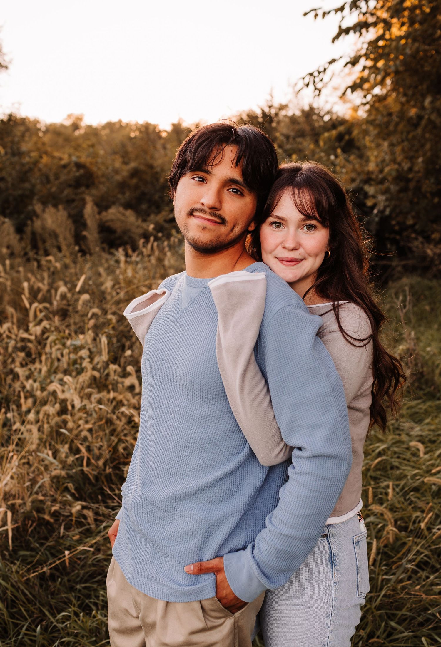 A young couple embraces in a golden field at sunset, wearing casual blue and beige tops, smiling warmly.