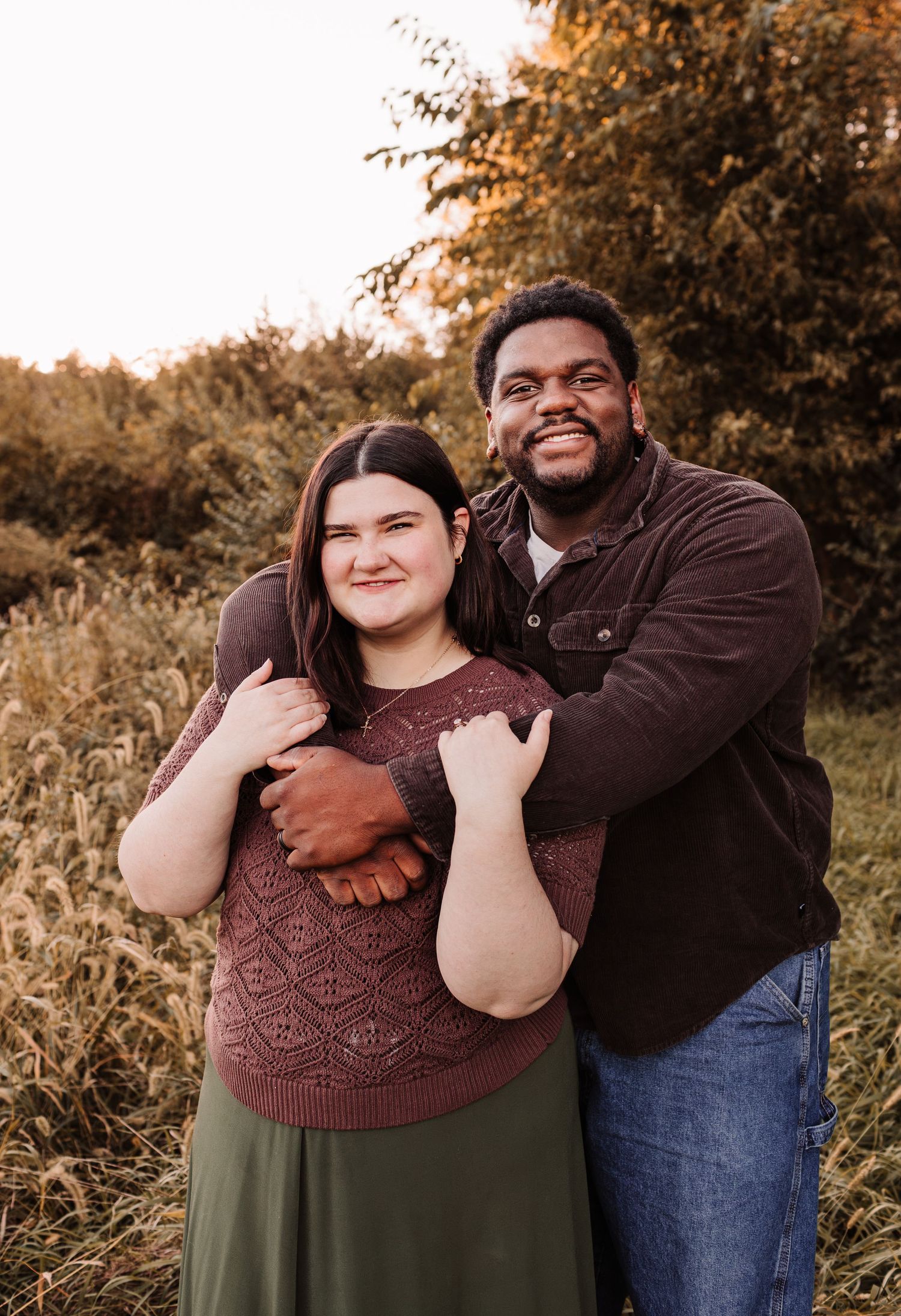 Couple embracing outdoors in autumn field, man in brown shirt hugging woman in mauve sweater and green skirt.
