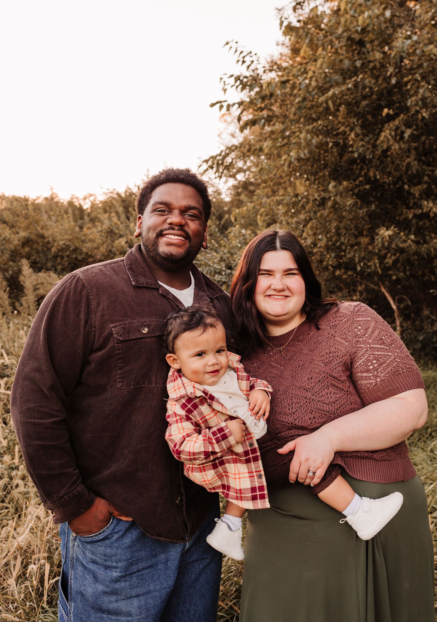 A smiling family of three poses outdoors, with parents holding their baby dressed in plaid, surrounded by lush greenery.