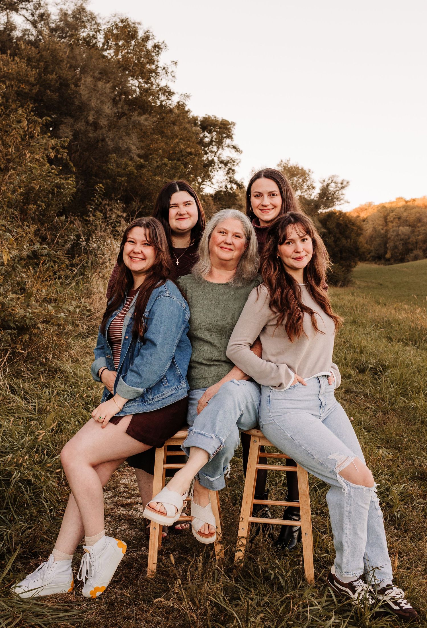 A woman and four young women pose together outdoors in a field during golden hour for a family portrait.