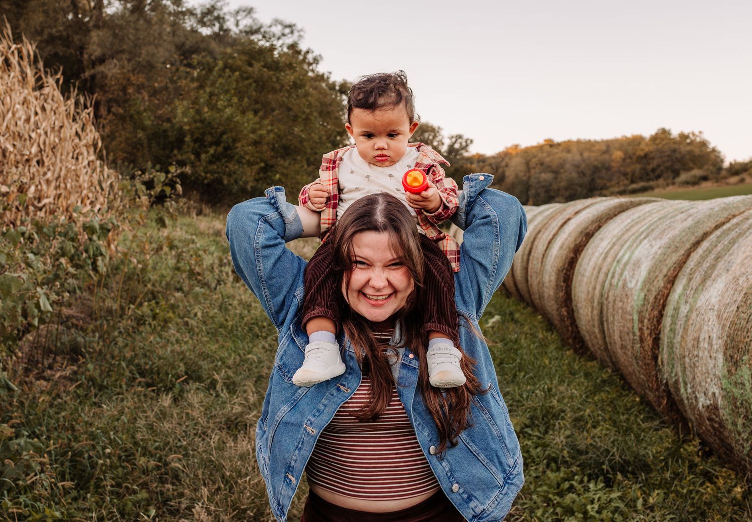 A smiling young woman carries a baby on her shoulders in a sunny farm field with hay bales and green trees.