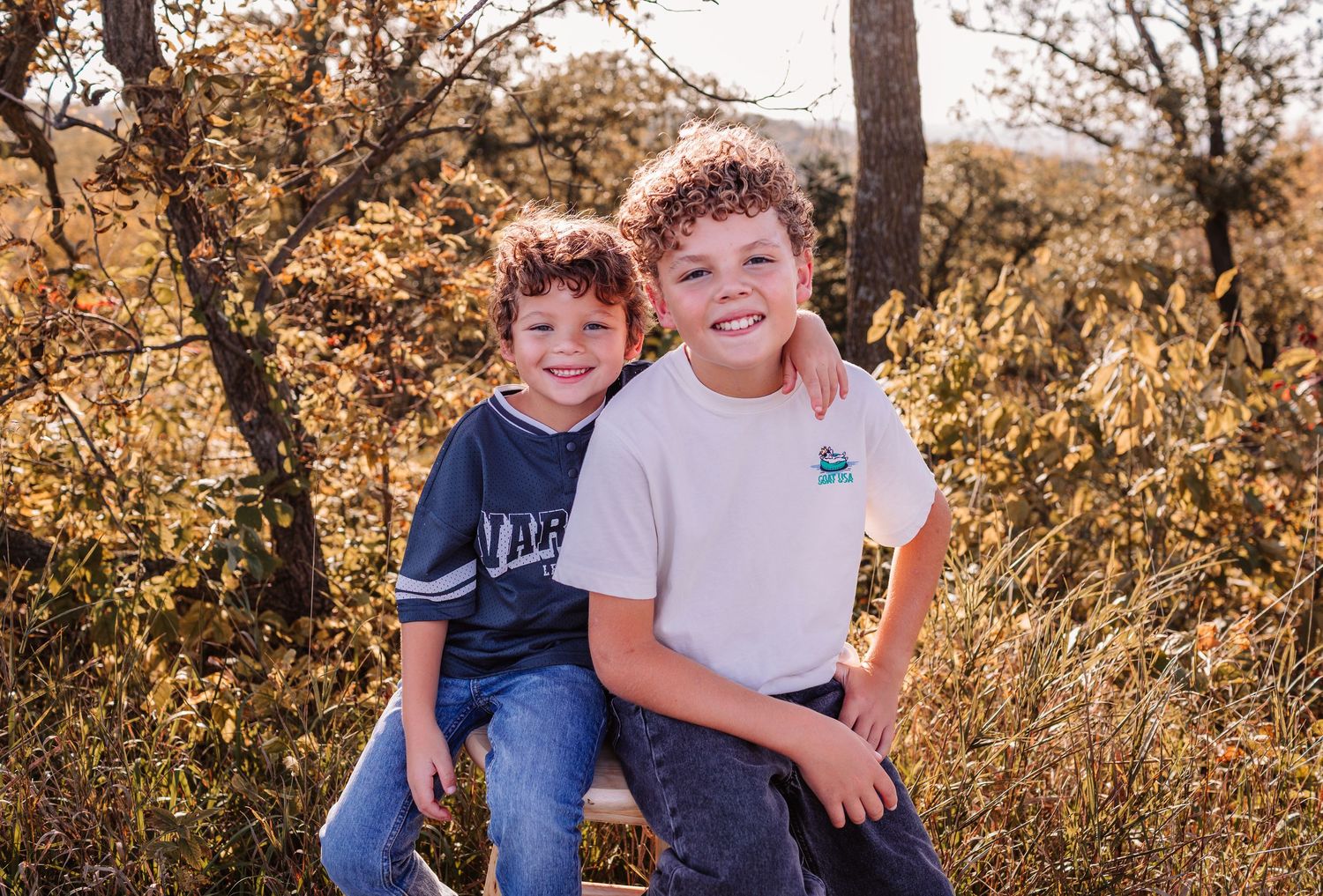 Two smiling boys pose together outdoors among autumn foliage trees in a warm fall nature setting.