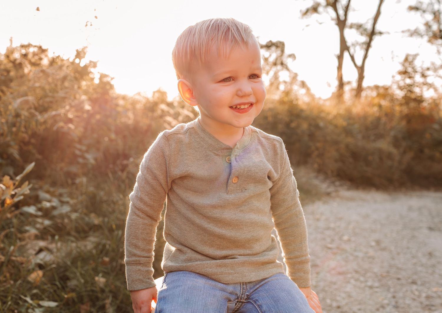 A happy toddler boy with blonde hair sits outdoors in golden sunset light, wearing a beige henley shirt and blue jeans.