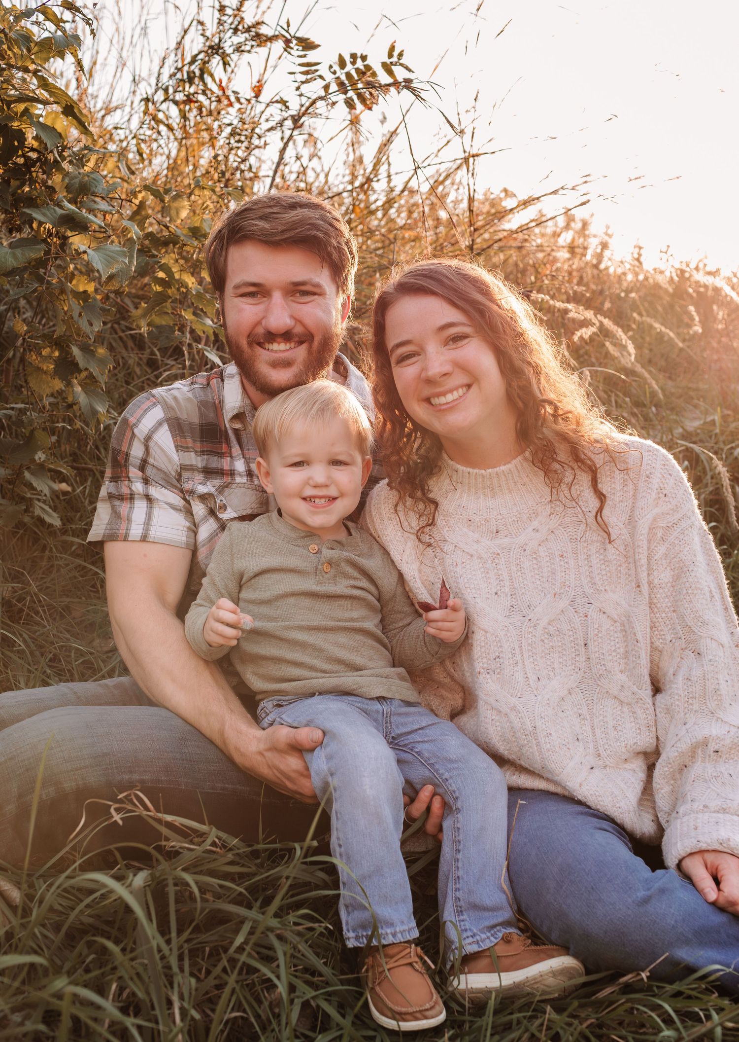 Happy young family with toddler posing outdoors in golden hour light among tall autumn grasses.