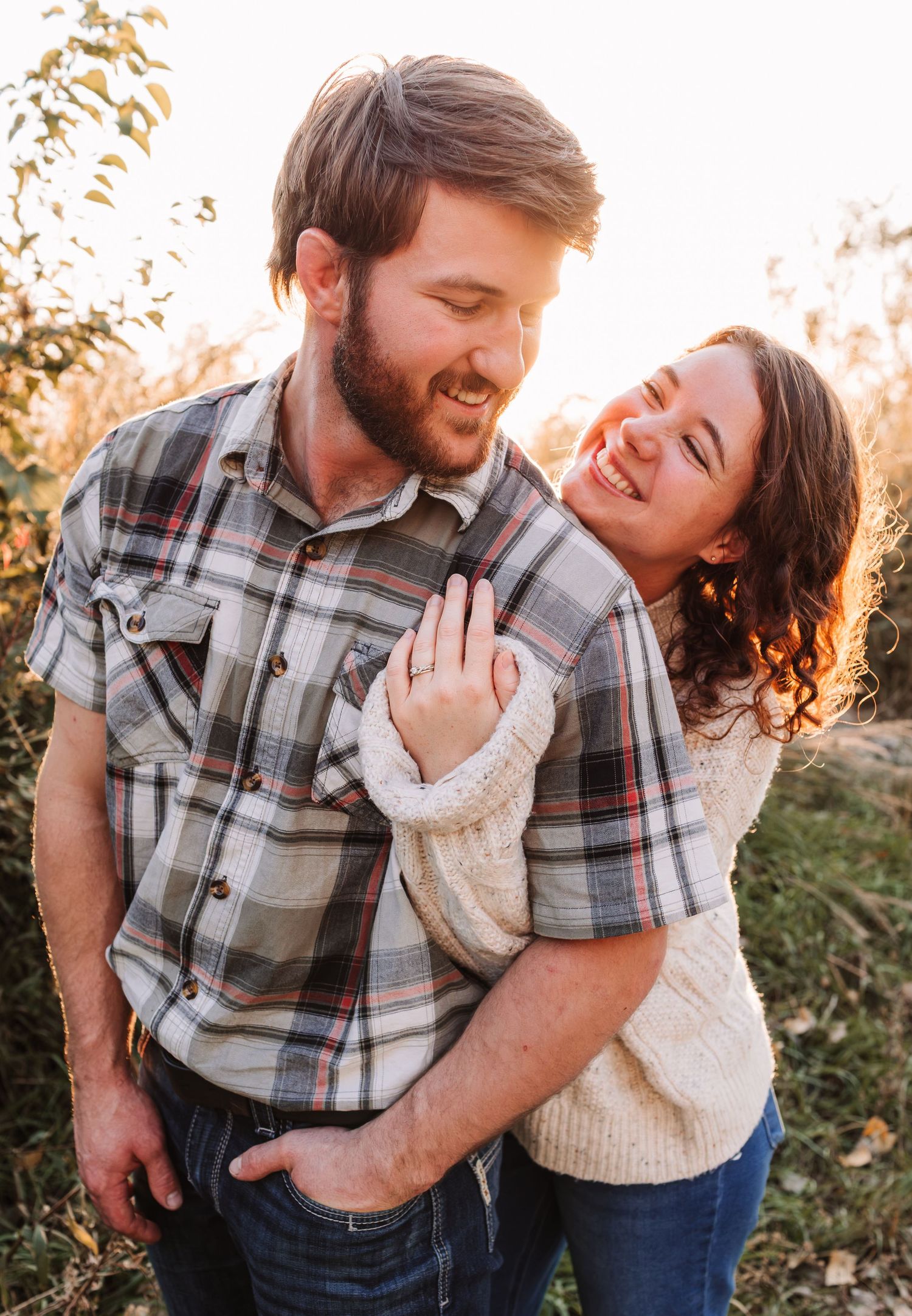 Happy couple embracing outdoors in golden autumn light, man in plaid shirt, woman in cream sweater smiling joyfully.