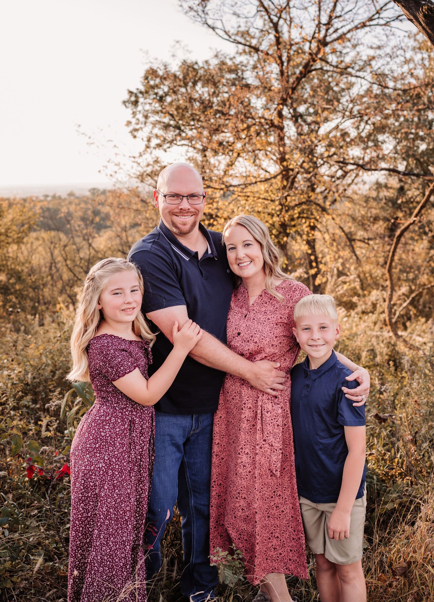 Family of four smiling outdoors during golden hour, parents embracing two children in coordinating navy and burgundy outfits.