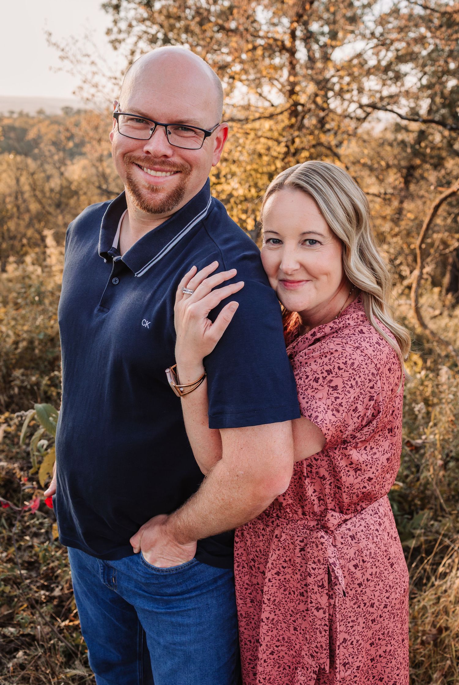 Couple posing outdoors at golden hour, man in navy polo and jeans, woman in pink floral dress, autumn foliage background.