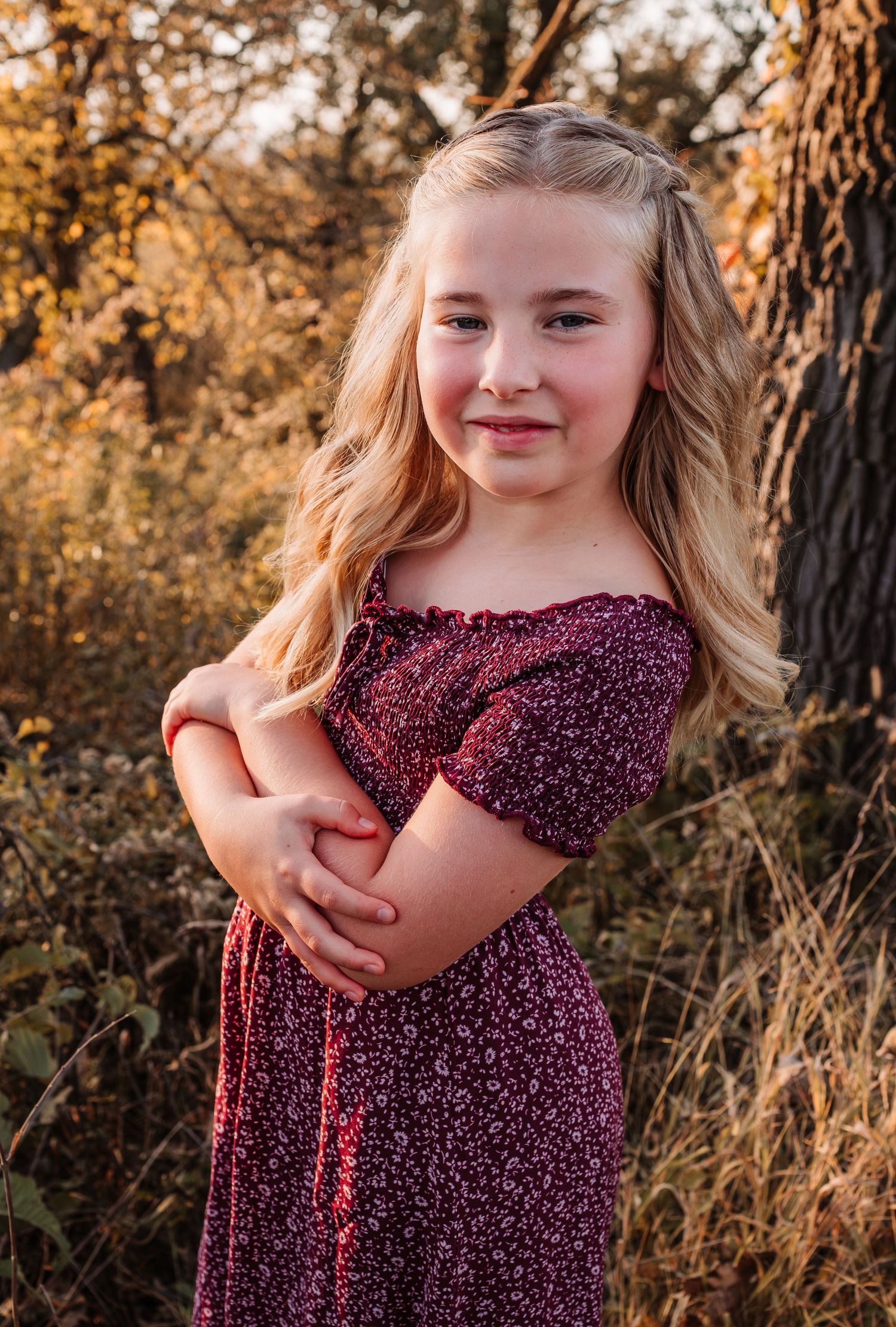 Young blonde girl in a floral burgundy dress posing outdoors among autumn trees in golden light.