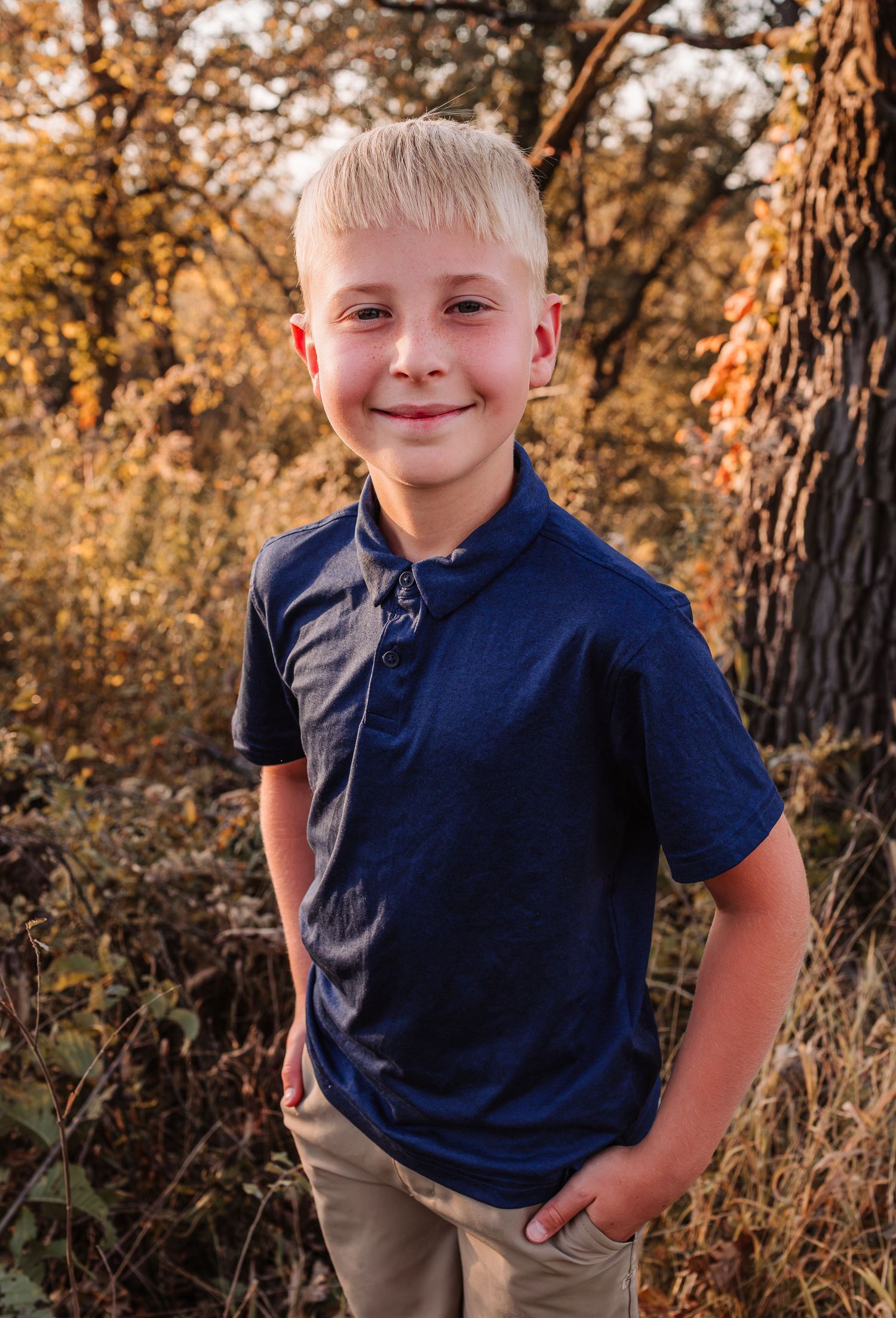 Smiling blonde boy in navy polo shirt posing outdoors among autumn foliage trees.