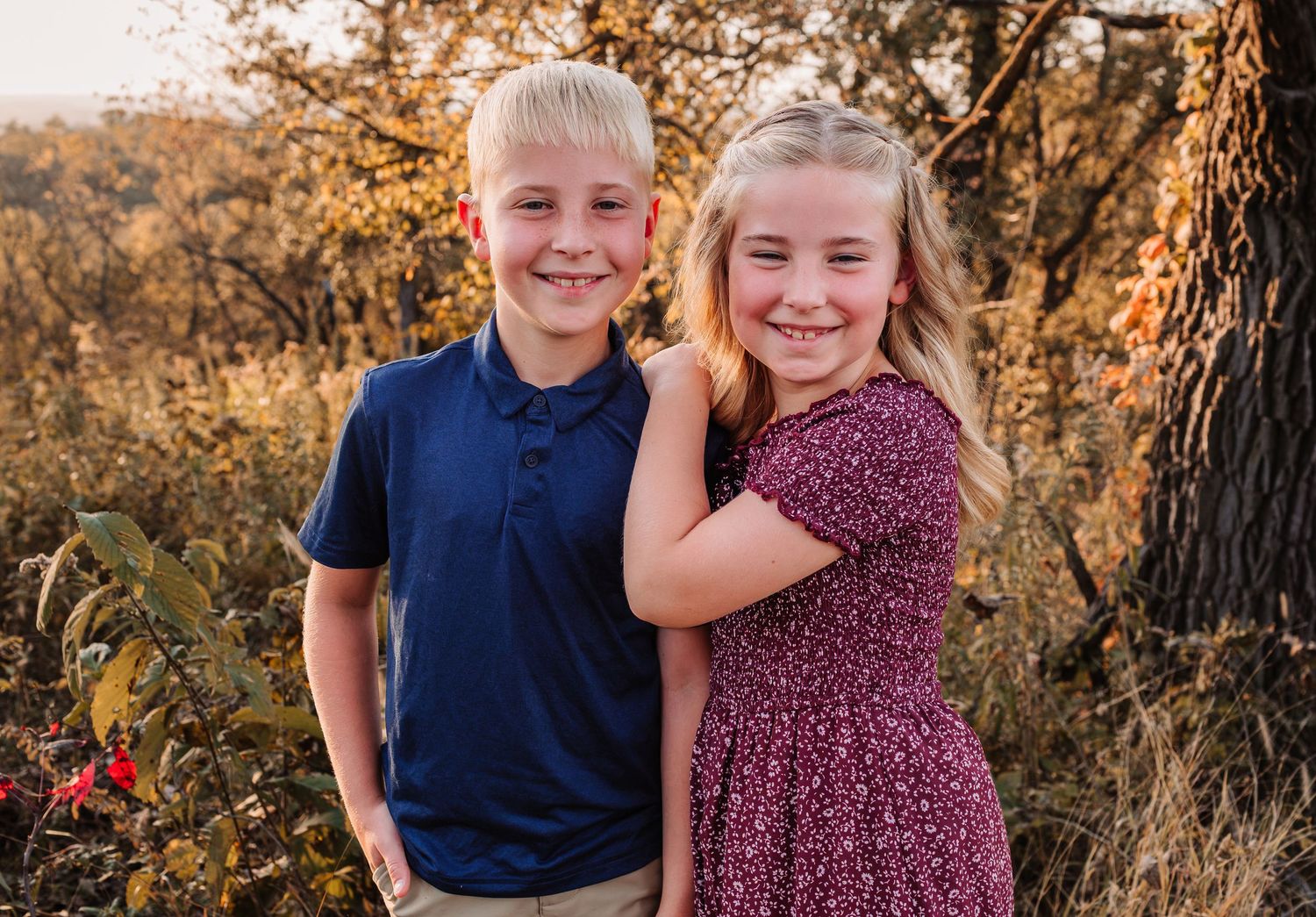 Two smiling children pose outdoors in autumn, boy in navy polo and girl in floral dress, surrounded by golden fall foliage.