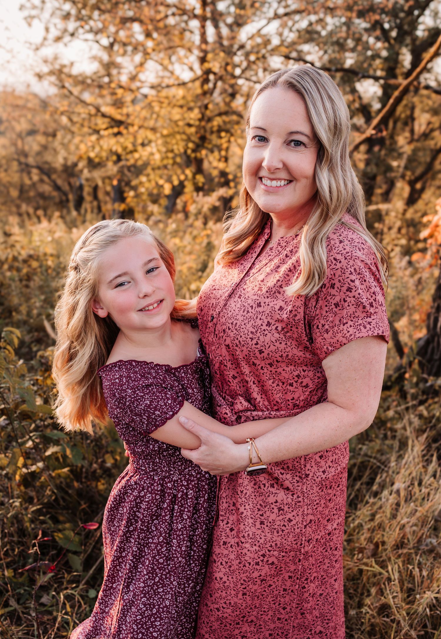 A smiling woman in a pink dress poses with a young blonde girl in a purple dress outdoors in autumn.