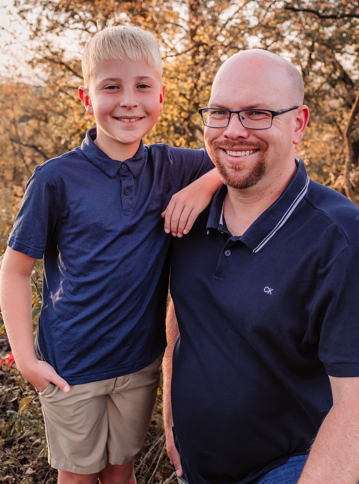 A smiling man and young blonde boy wearing matching navy polo shirts pose together outdoors in warm autumn light.
