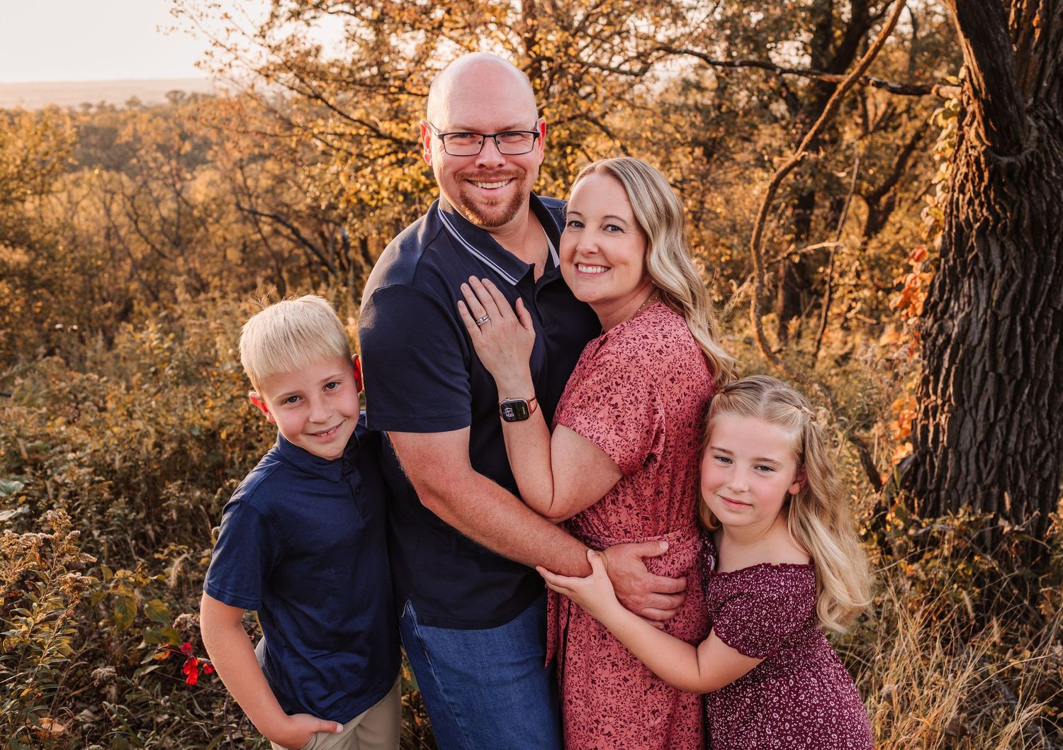 Family of four smiling outdoors during golden hour, parents embracing two children among autumn foliage.