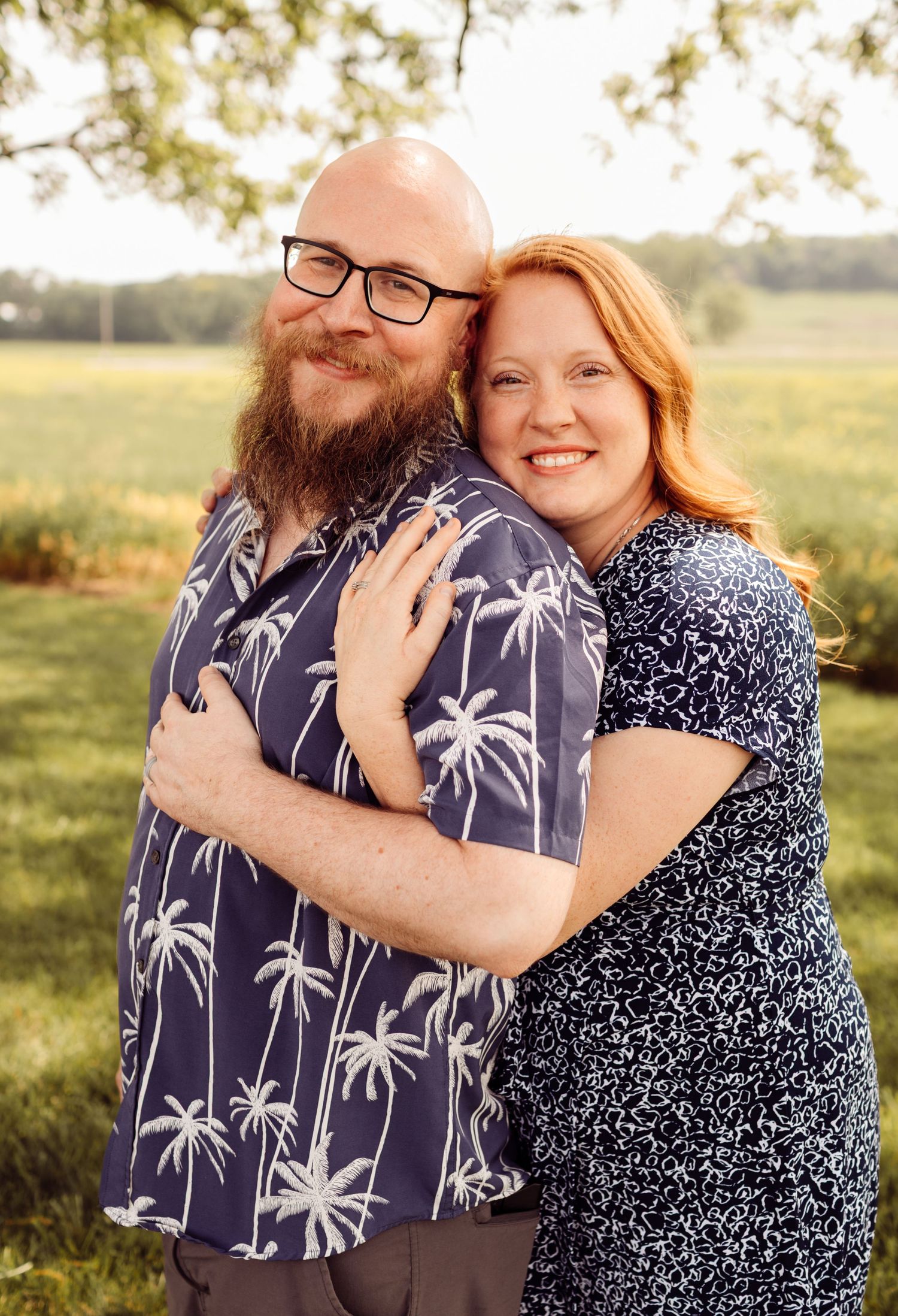 Couple embraces outdoors at golden hour, man in palm tree shirt, woman in blue floral dress.