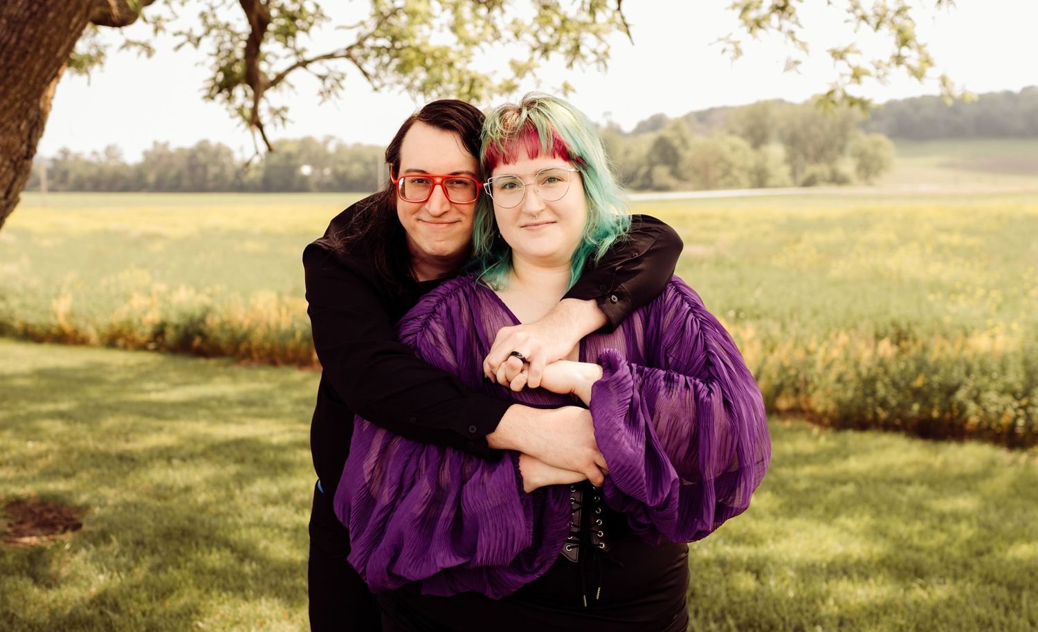 Two women hug warmly outdoors, one in purple jacket with teal hair highlights, golden field behind.