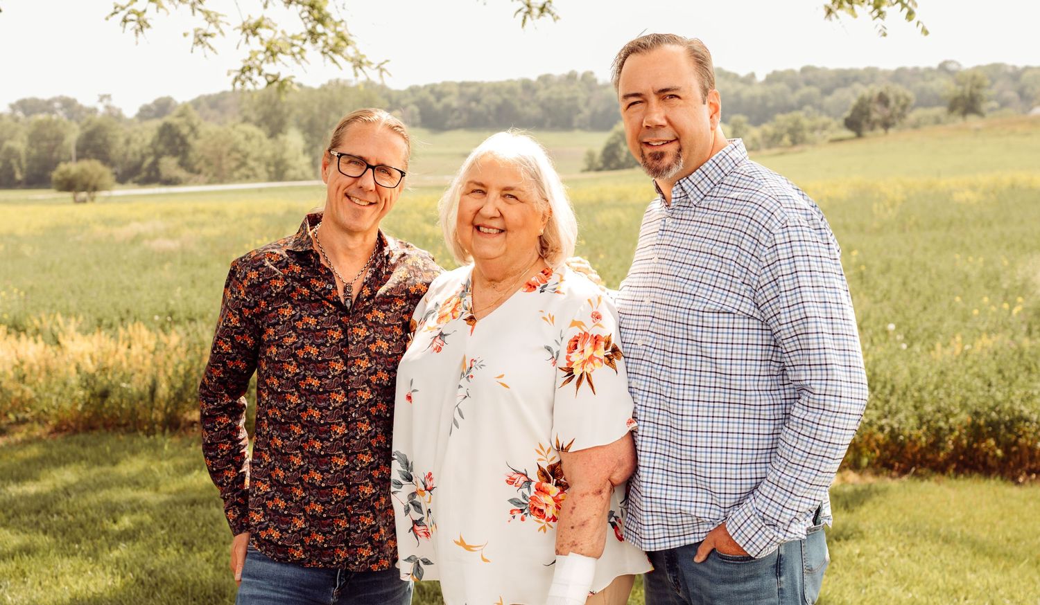 Two men and a woman smile together outdoors in a sunny green field family portrait.