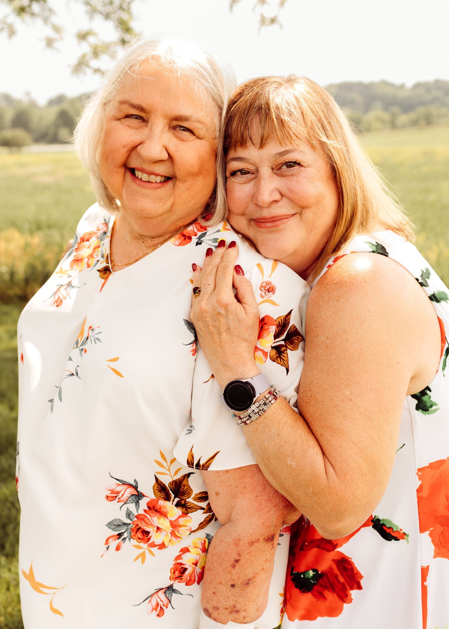 Two women in matching floral dresses embrace warmly, smiling together in an outdoor summer setting.