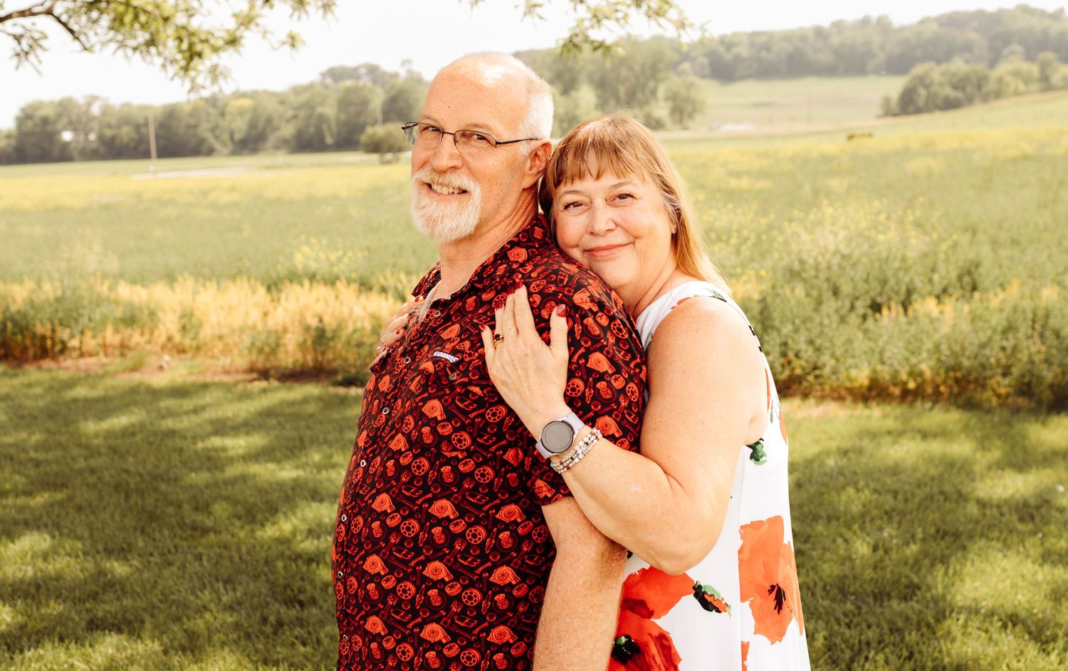 Older couple poses affectionately in a sunny green field, man in red patterned shirt.