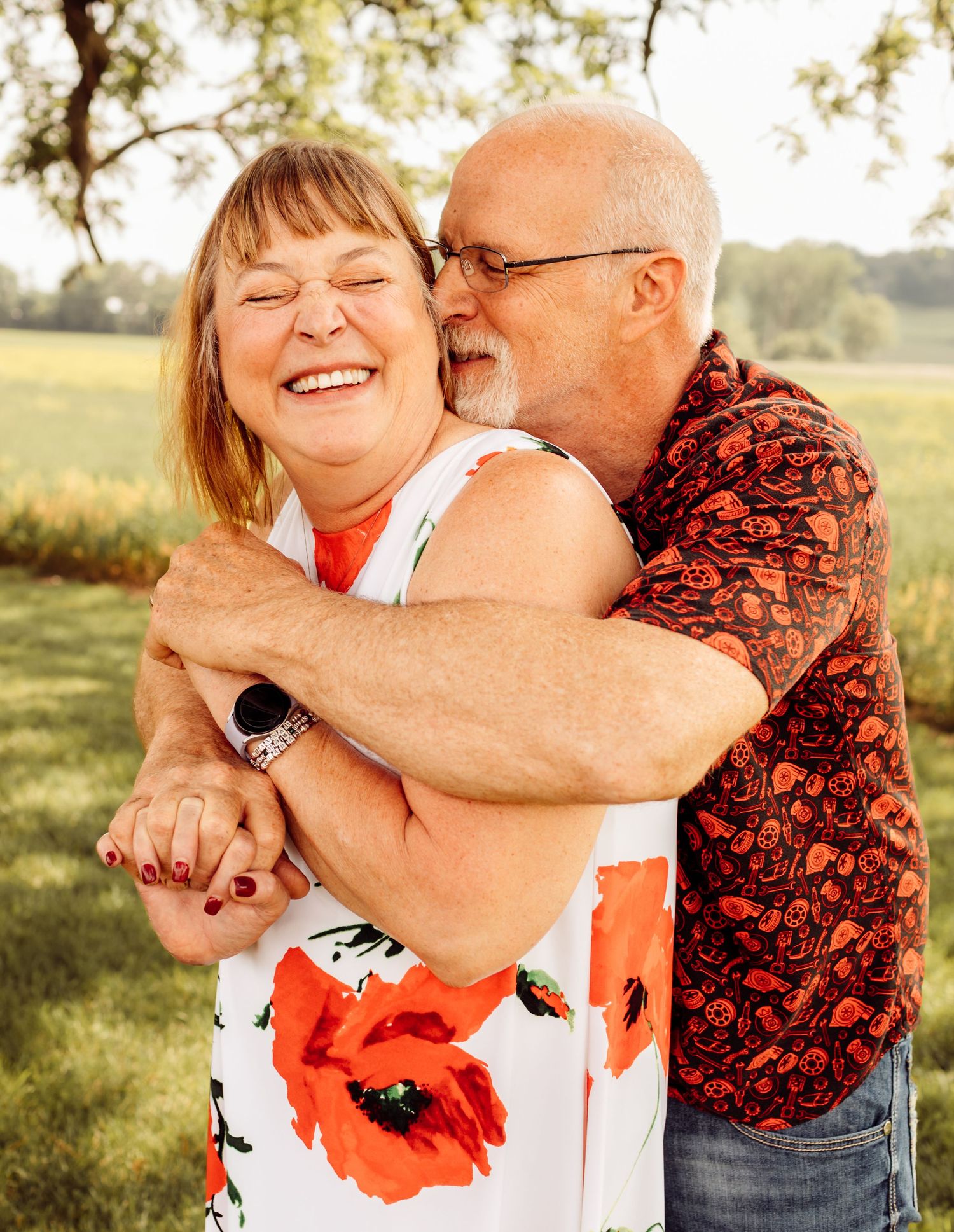 Joyful older couple embraces outdoors, woman laughing as man kisses her cheek in golden sunlight.