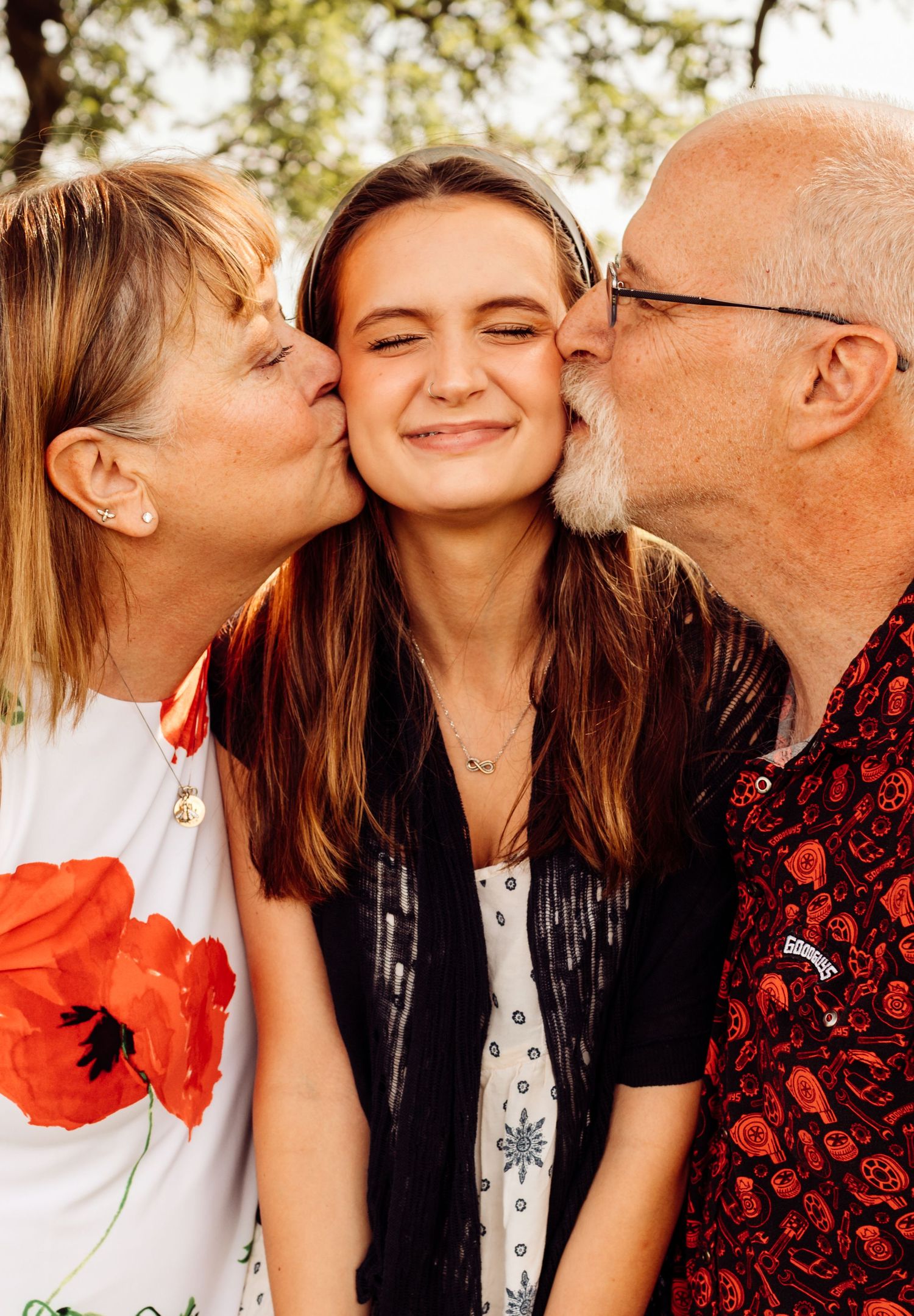 Teenage girl smiles as grandparents kiss her cheeks simultaneously in a warm outdoor portrait.