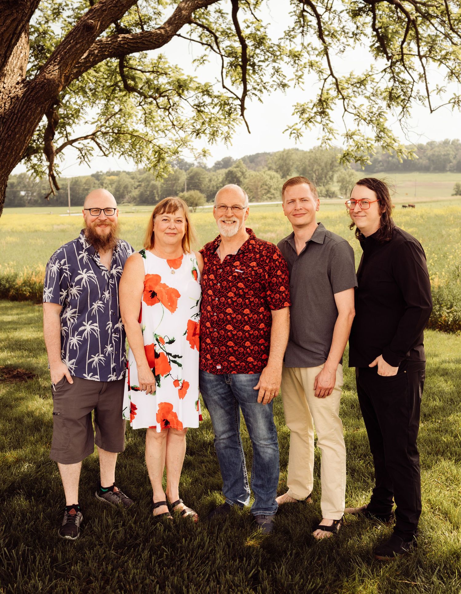 Family of five smiling outdoors under a tree in a green field during golden hour.
