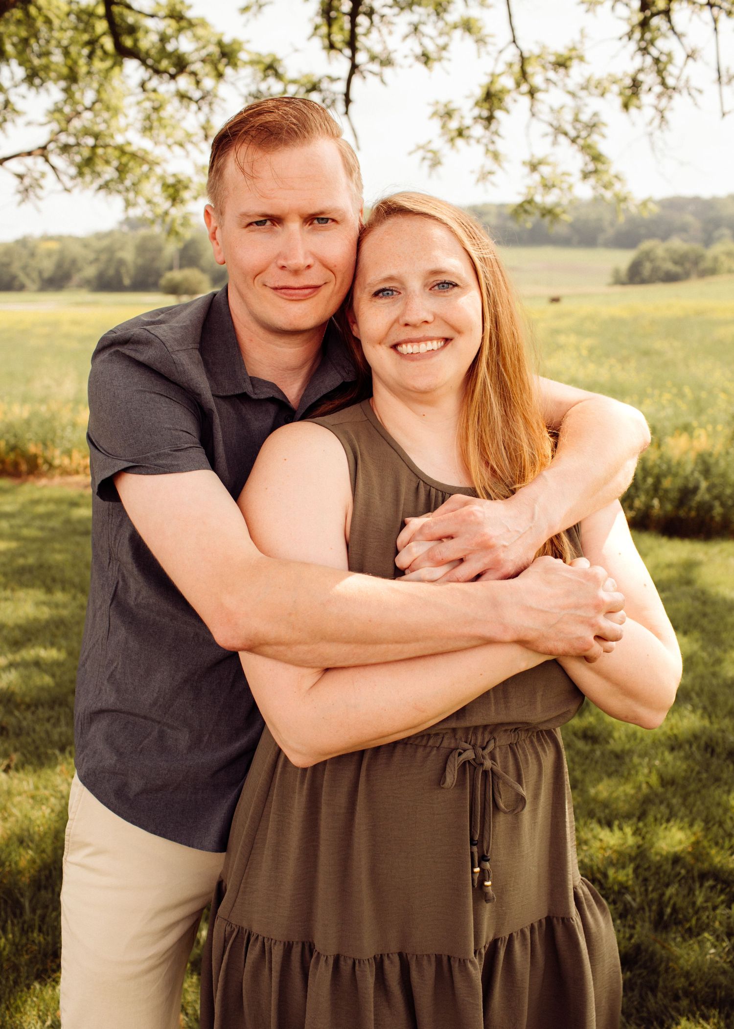 Couple embracing and smiling together outdoors in a green field during golden hour portrait session.
