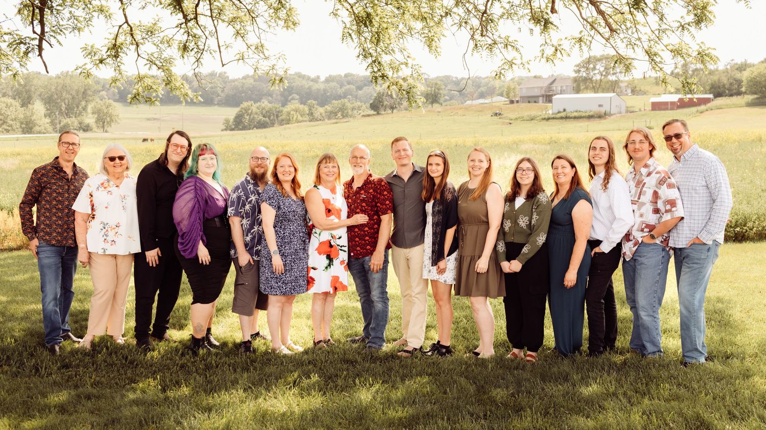 Large extended family group portrait outdoors under a tree in a green field at sunset.