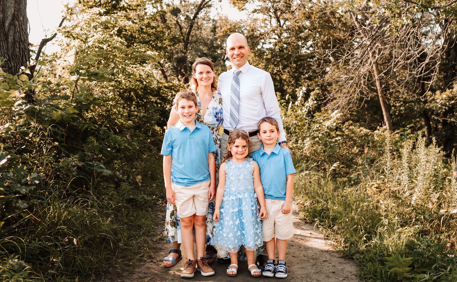 Happy family of five posing on a nature trail surrounded by lush green trees in coordinating blue outfits.