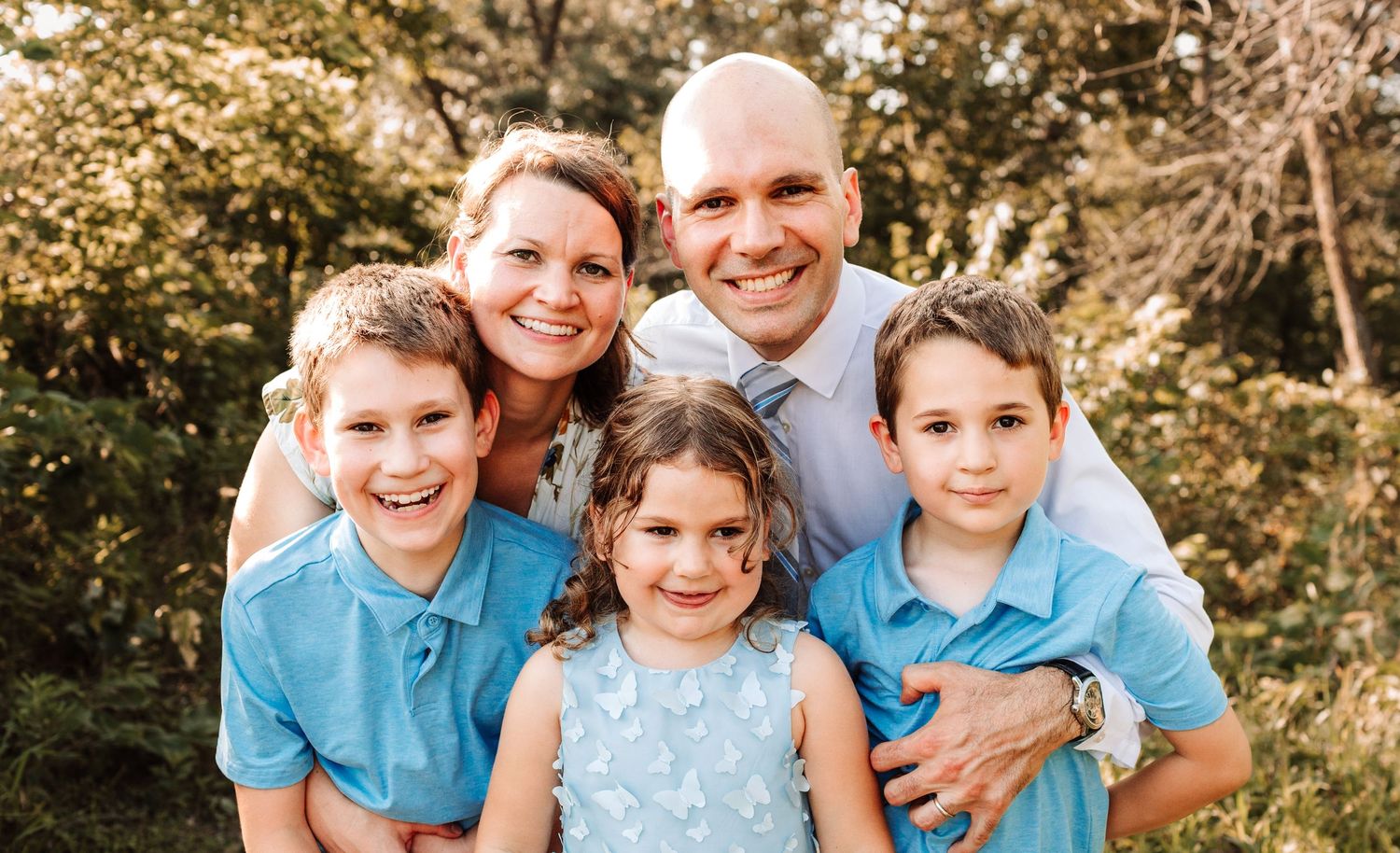 Smiling family portrait of parents with three young children wearing coordinating blue outfits outdoors.