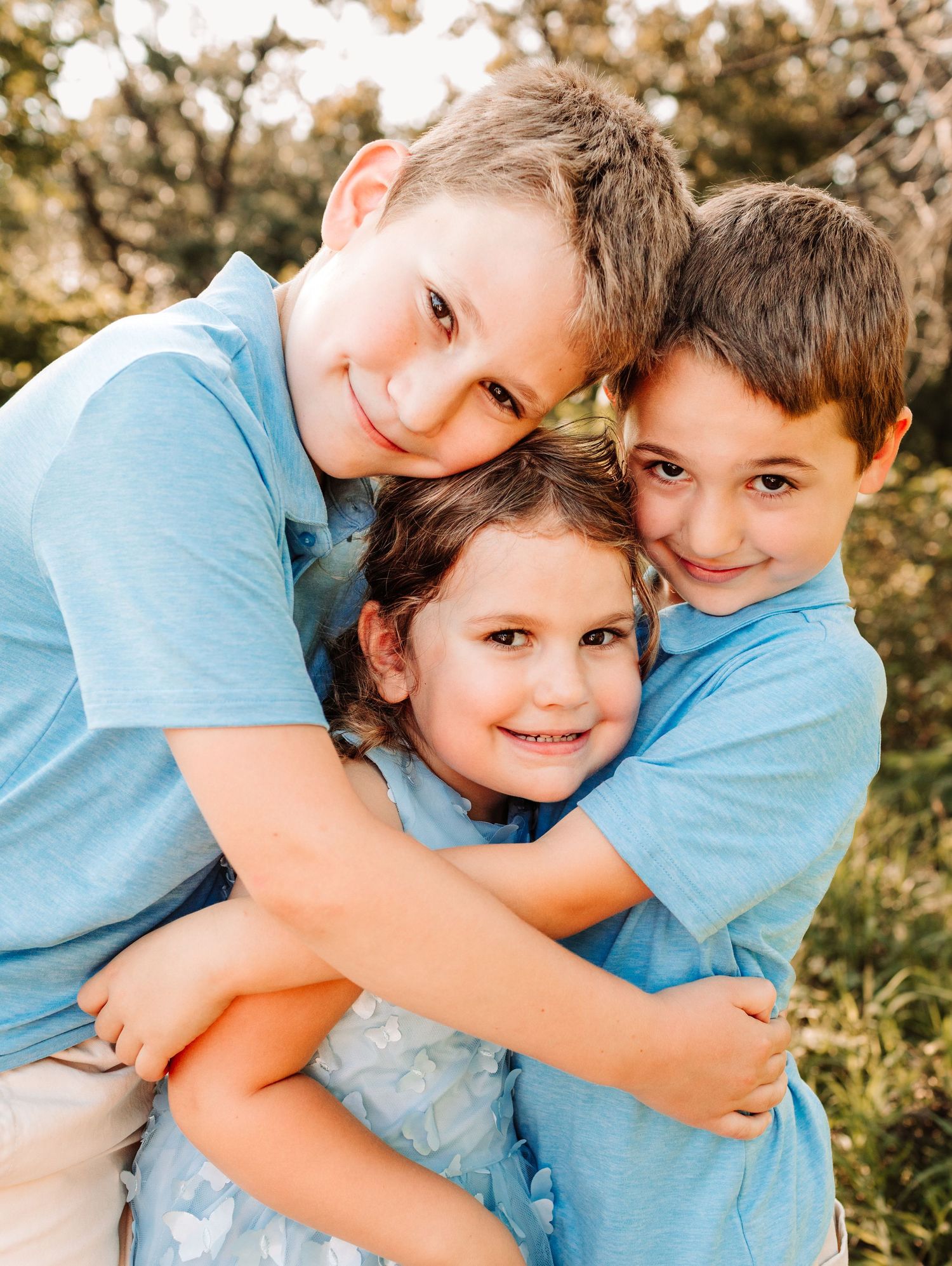 Three siblings in blue outfits hugging and smiling together in a warm outdoor portrait session.