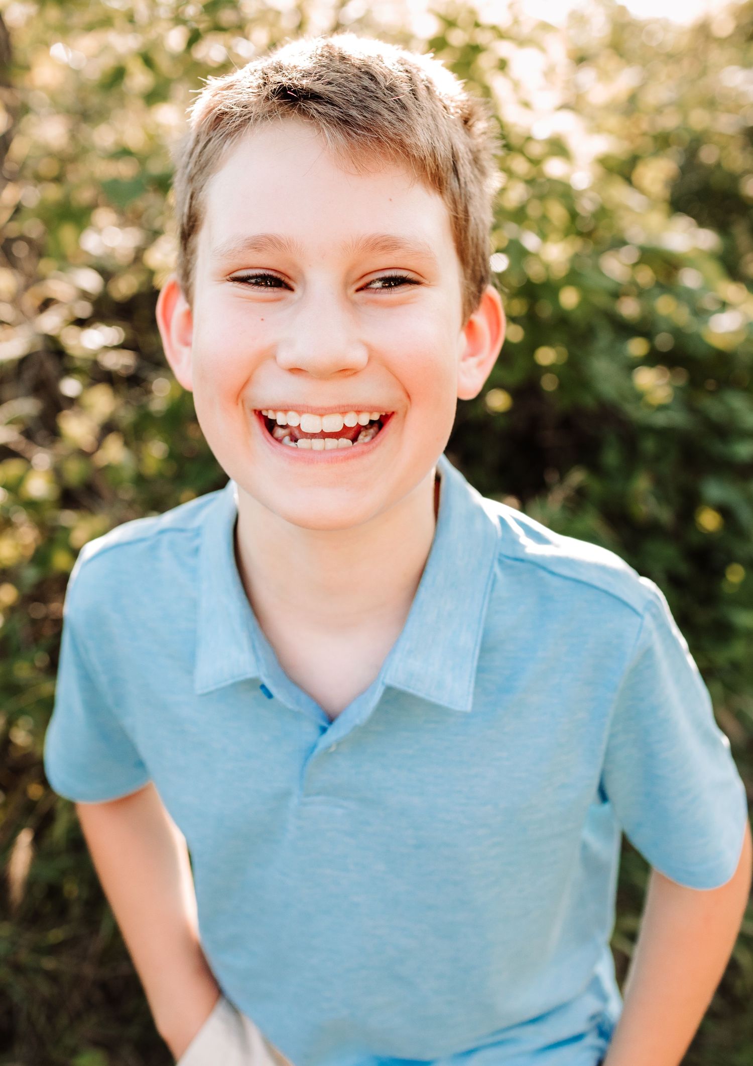 Cheerful young boy with short blonde hair smiling brightly in a light blue polo shirt outdoors.